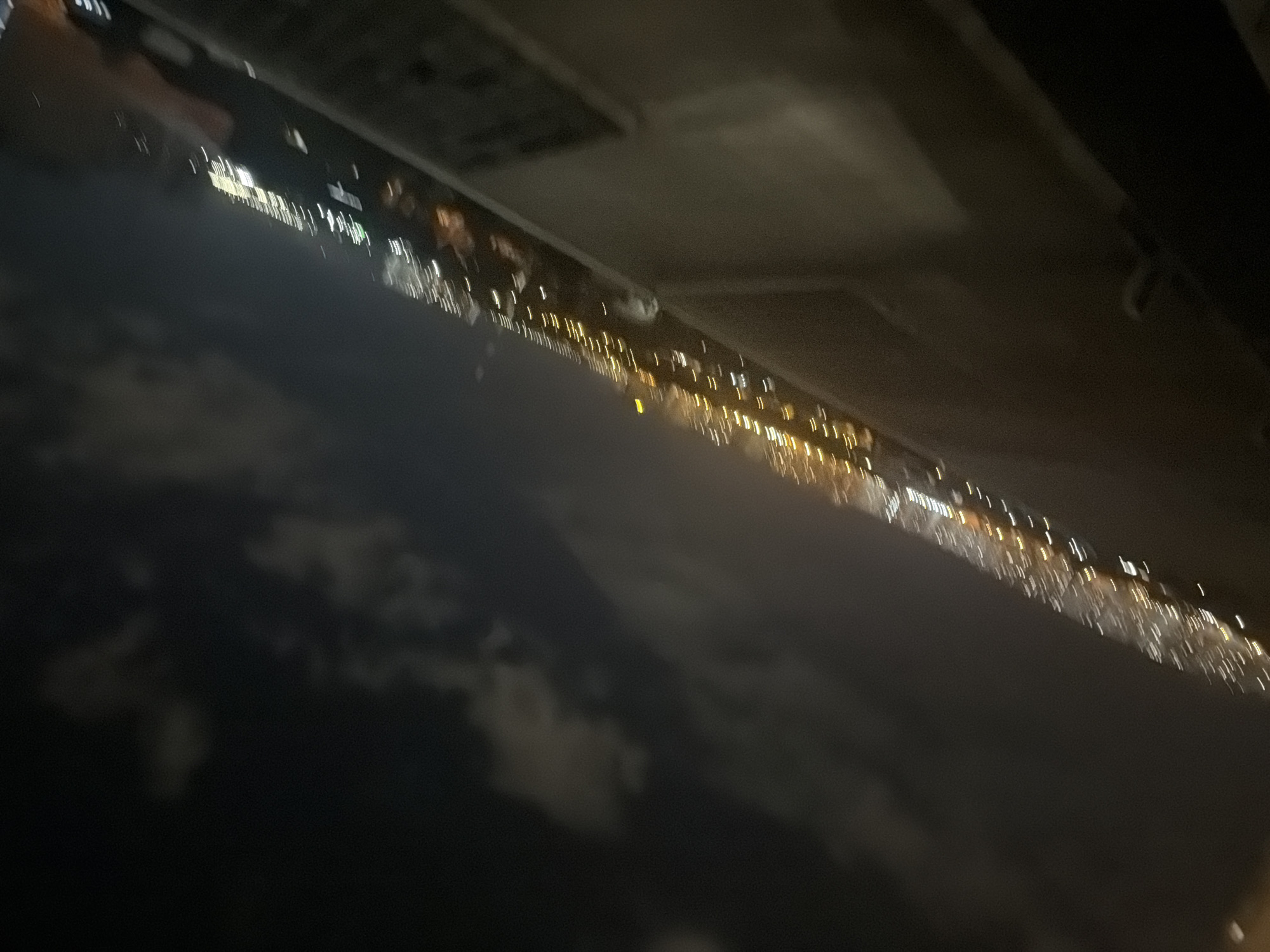 This is a blurry, night-time aerial view of a city, likely taken from an airplane window. The image is dominated by streaks of light representing city lights, suggesting a densely populated urban area. The lights are blurred due to motion, indicating the camera was moving, possibly during flight. The sky above is dark with some faint cloud formations visible. The foreground is obscured by a dark, angled surface, which could be part of the aircraft or a balcony. The city lights appear to be stretching along a line, perhaps following a river or a major road. The specific location in Kyiv, Ukraine, cannot be definitively determined due to the lack of clear landmarks and the image's blurriness.