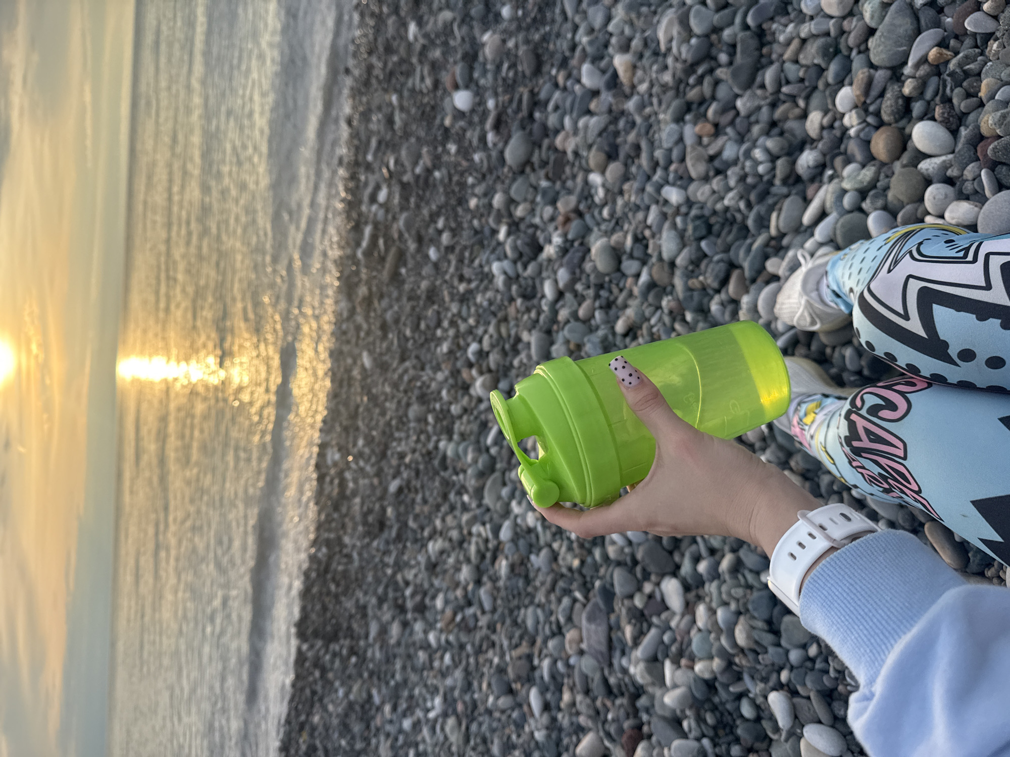 The image captures a person seated on a pebble beach, holding a green plastic water bottle. The setting sun is visible over the sea. The person is wearing patterned leggings and white sneakers. A white smartwatch is on their wrist. The beach is composed of numerous small, smooth, grey pebbles. This photo could be taken in Batumi, Georgia.