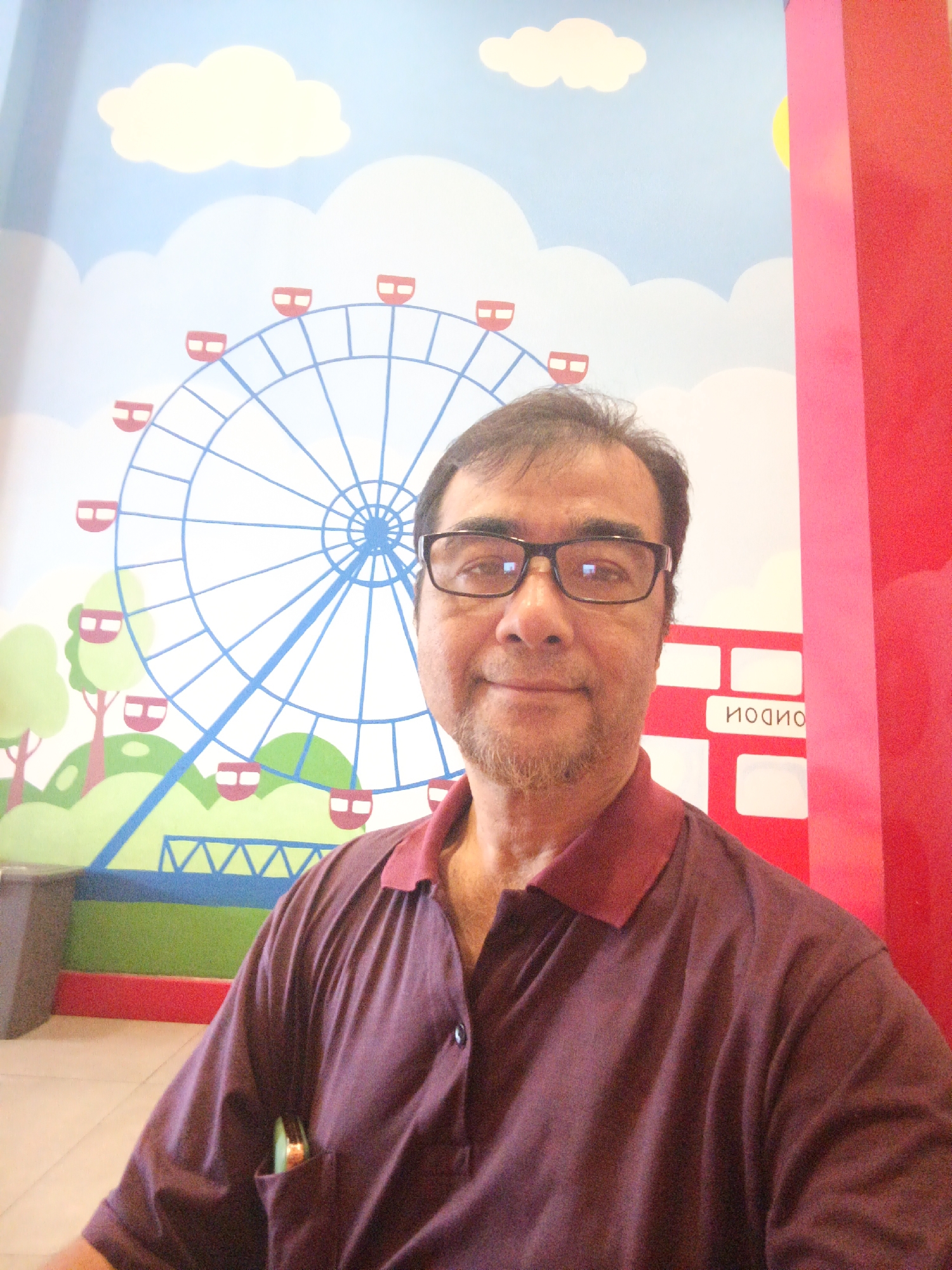 A middle-aged man wearing glasses and a maroon polo shirt is taking a selfie in front of a mural. The mural depicts a bright blue sky with white clouds, a large Ferris wheel with red gondolas, and green hills with cartoonish trees. To the right of the mural, there is a red and pink vertical structure. In the background, part of a red bus with the word "LONDON" written in white letters is visible on the mural. The man has a goatee and appears to be smiling. He has a small, green object in his shirt pocket. The lighting suggests an indoor environment, possibly a public space like a mall or an amusement center. The image appears to be taken in Depok, Indonesia.
