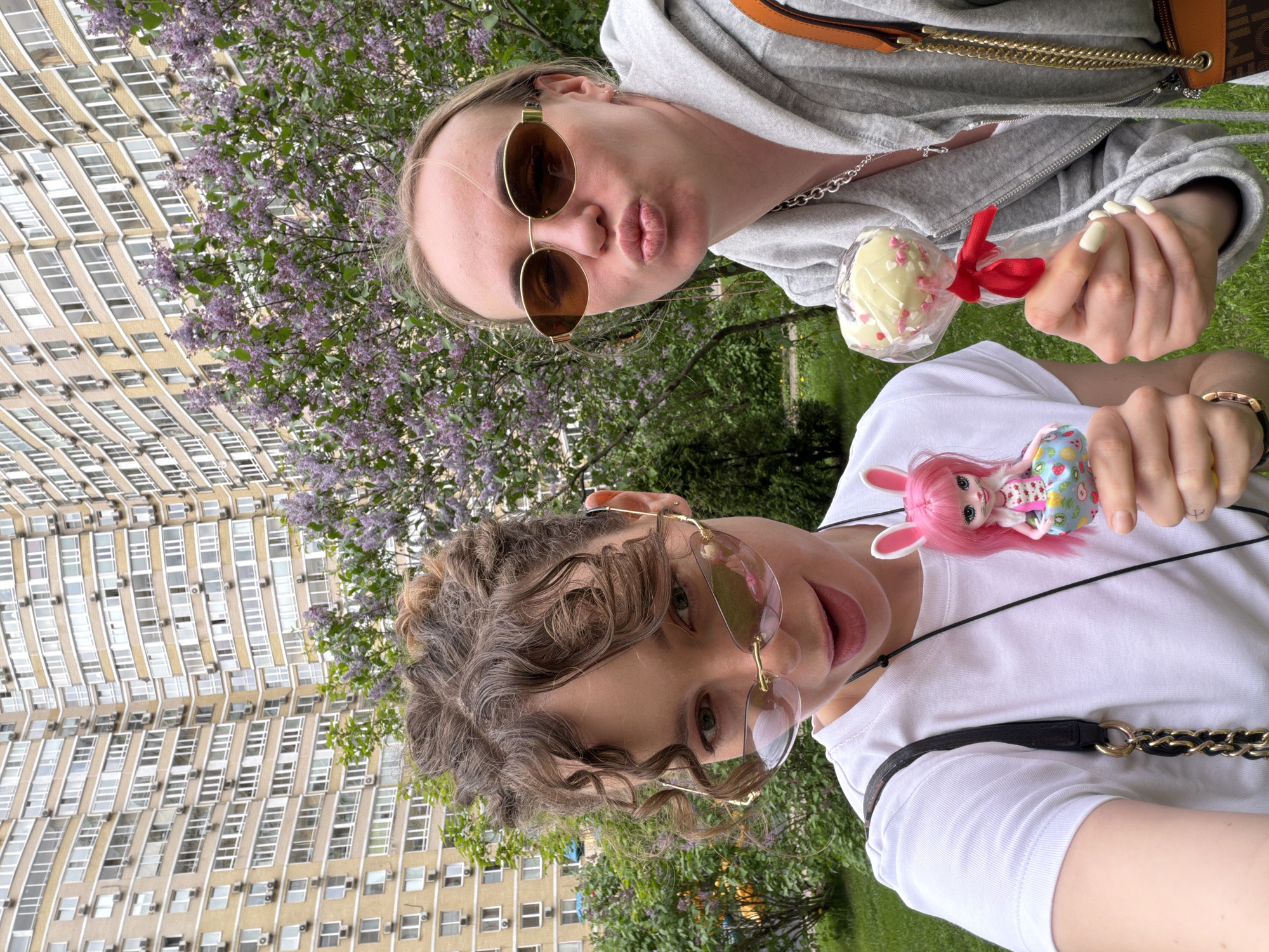Two individuals are visible in the foreground of a photograph, seemingly captured from a low-angle selfie perspective. The person at the top is wearing a grey hoodie, gold-framed sunglasses and is puckering their lips. They are holding a lollipop with a red ribbon. The second person at the bottom is wearing a white shirt, gold-framed glasses, and holding a small doll with pink hair and bunny ears. In the background, there is a tall building with multiple windows, partially obscured by green foliage and purple lilac bushes. Based on the architecture, the location is likely Moscow, Russia.