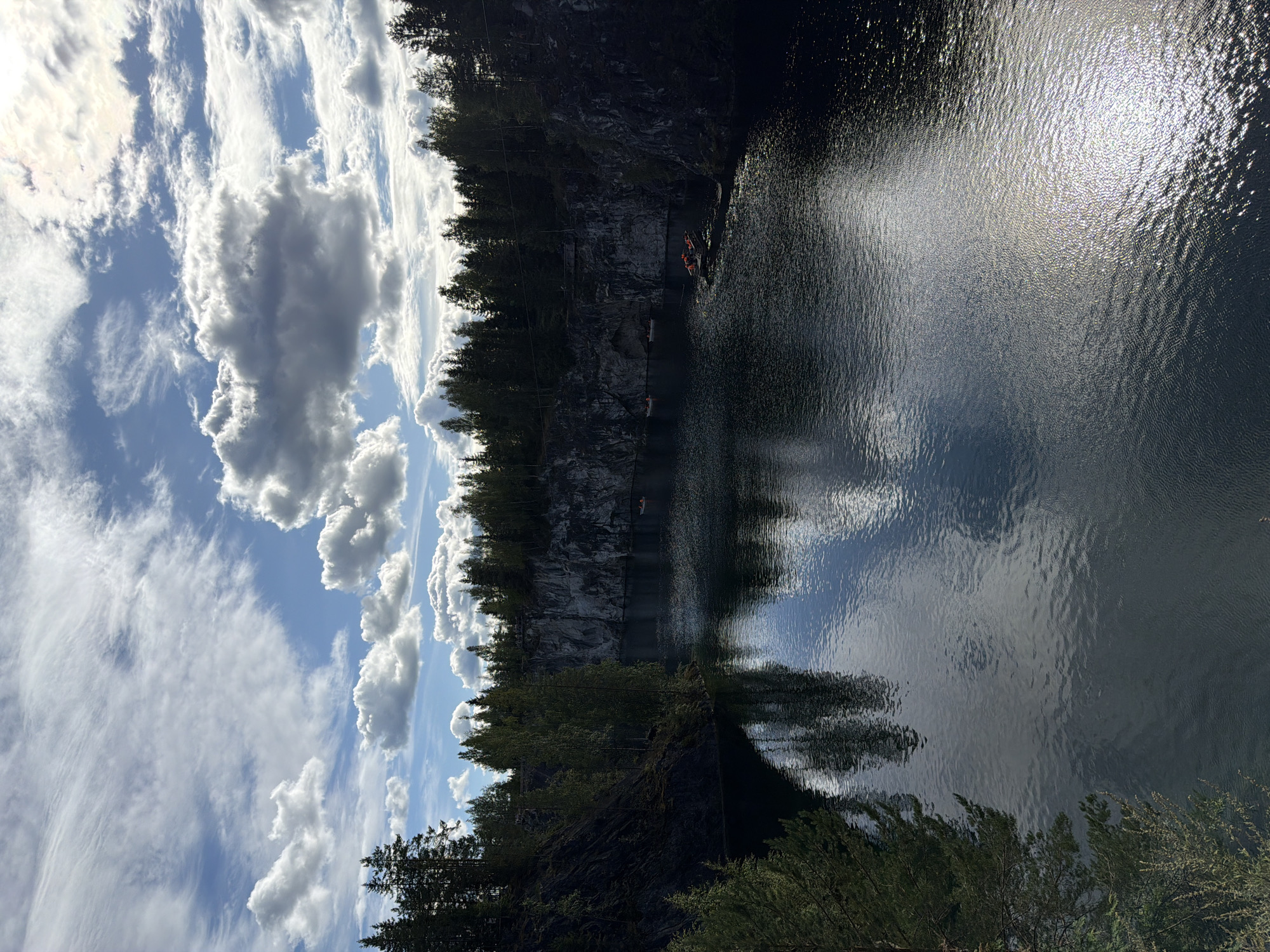 A vertical, high-angle shot captures a quarry lake surrounded by steep, rocky cliffs and dense evergreen trees under a partly cloudy sky. The lake's dark, rippling surface reflects the sky, trees, and cliffs, with sunlight glinting off the water in the lower right corner. Several small boats, some with people wearing orange life vests, are visible scattered across the lake, suggesting recreational activity. The quarry walls are rugged and show layers of rock, with vegetation growing at the top of the cliffs and on the slopes. The sky is a mix of bright blue patches and dramatic, shadowed clouds, with streaks of sunlight breaking through. The overall atmosphere is serene, showcasing a natural landscape transformed by past industrial activity.