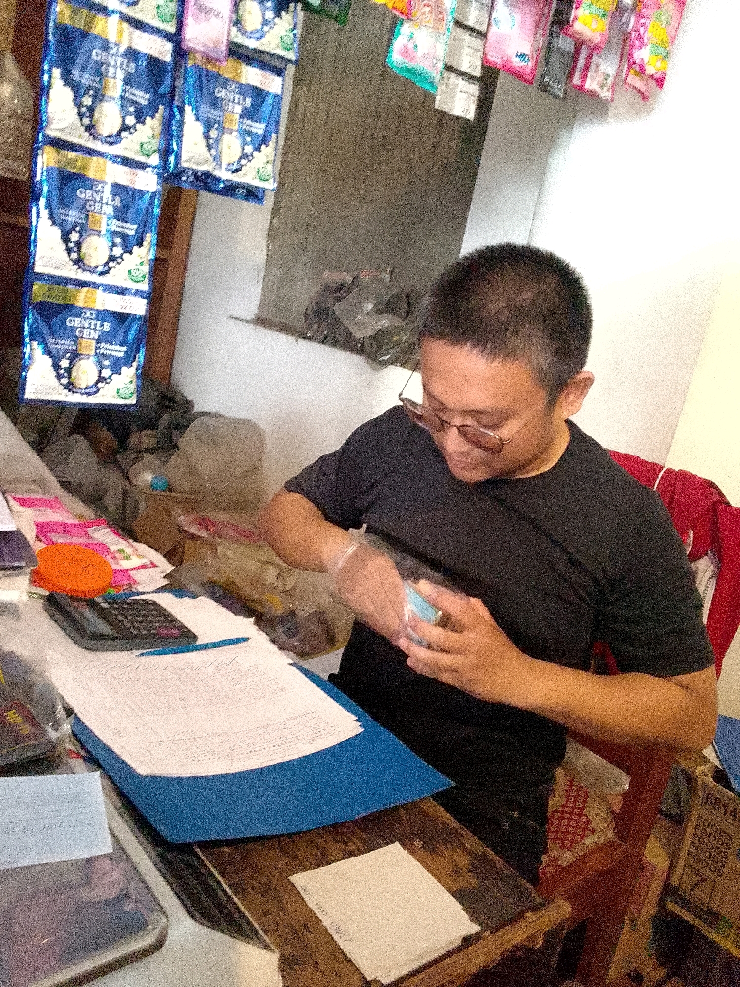 A person is seated at a wooden table in what appears to be a small shop or storage area in Cimahi, Indonesia. The person, wearing glasses and a black t-shirt, is holding a small, clear plastic bag in their gloved hands, examining its contents. On the table in front of them are various items: a calculator, a stack of papers with handwritten text, a blue folder, a small orange object, and other unidentifiable items scattered around. Behind the person, packets of "GENTLE GEN" powdered product are hanging on the wall, along with other items in colorful packaging. The background shows a textured wall and some cluttered shelves. The overall lighting suggests an indoor setting, possibly with natural light filtering in.