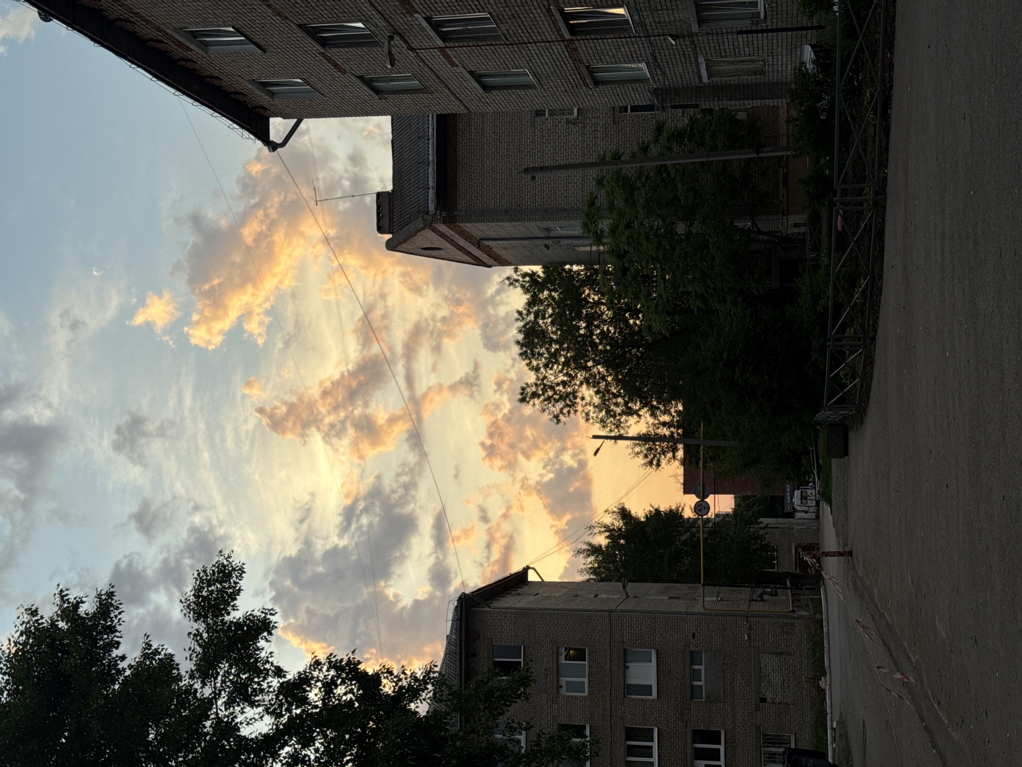 This image depicts a street scene in Perm, Russia. The view is angled upwards, capturing the sky filled with clouds illuminated by a sunset. Two multi-story brick buildings frame the sky, with trees partially obscuring the lower sections. A concrete sidewalk or street runs along the right side of the frame, with a fence visible along its edge. Further down the street, other buildings are visible in the distance. No people are visible in the image.