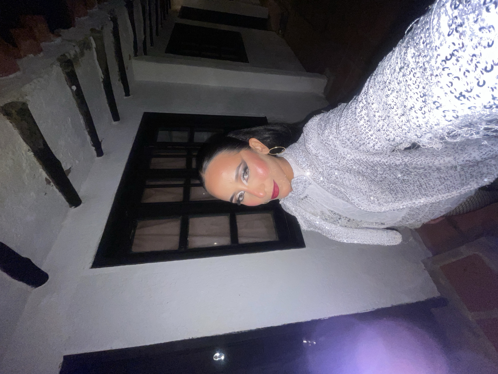 The image is a first-person, full-body, selfie-style shot of a woman in Porlamar, Venezuela. She is standing outdoors at night in front of a white building with dark windows and exposed wooden beams on the ceiling. She is wearing a sparkly white top and jacket. The woman has on dramatic makeup with a bold red lip, heavy white eyeshadow, and prominent blush. She is also wearing large gold hoop earrings and her dark hair is pulled back into a ponytail. The background shows a narrow walkway with a dark doorway on the left and more windows and an open doorway leading to a darker area on the right. There is a purple light source to the left of the woman.