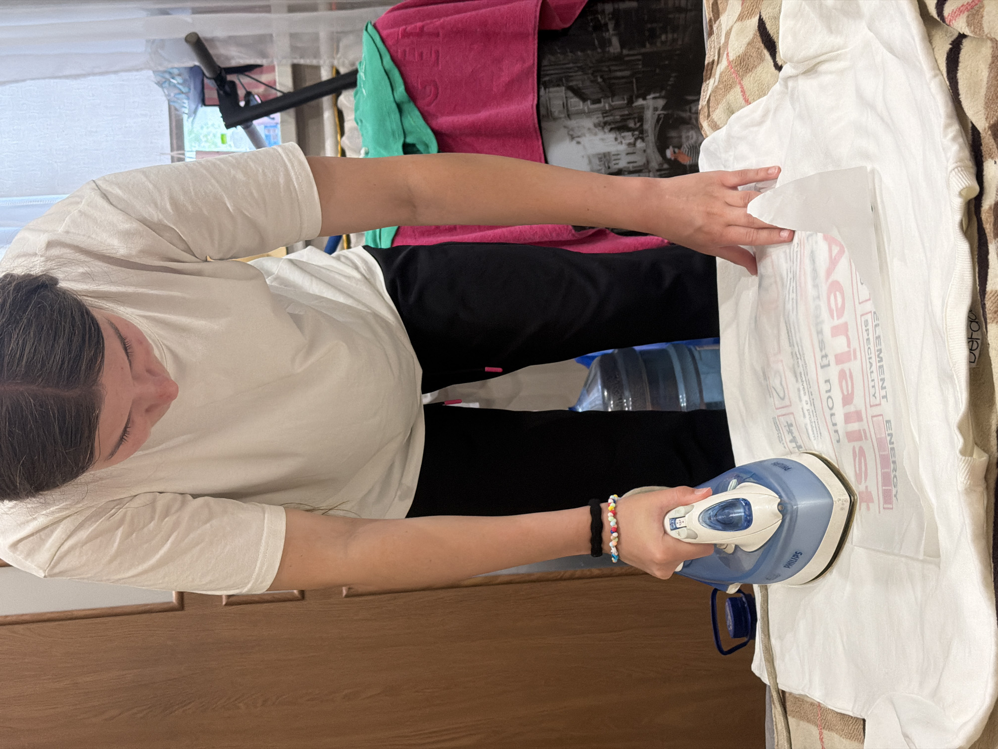 A young person is ironing a white t-shirt with a transfer graphic. The t-shirt is laid flat on a surface that appears to be a bedspread. The person is wearing a white t-shirt and black pants and has bracelets on their left wrist. The ironing is being done with a blue and white Philips iron. In the background, there is a large water cooler, a wooden closet, and a window with a scooter visible outside. The scene appears to be indoors, possibly in a residential setting. The presence of a water cooler suggests the location might be in the city of Dnipro, Ukraine, where these are commonly used.
