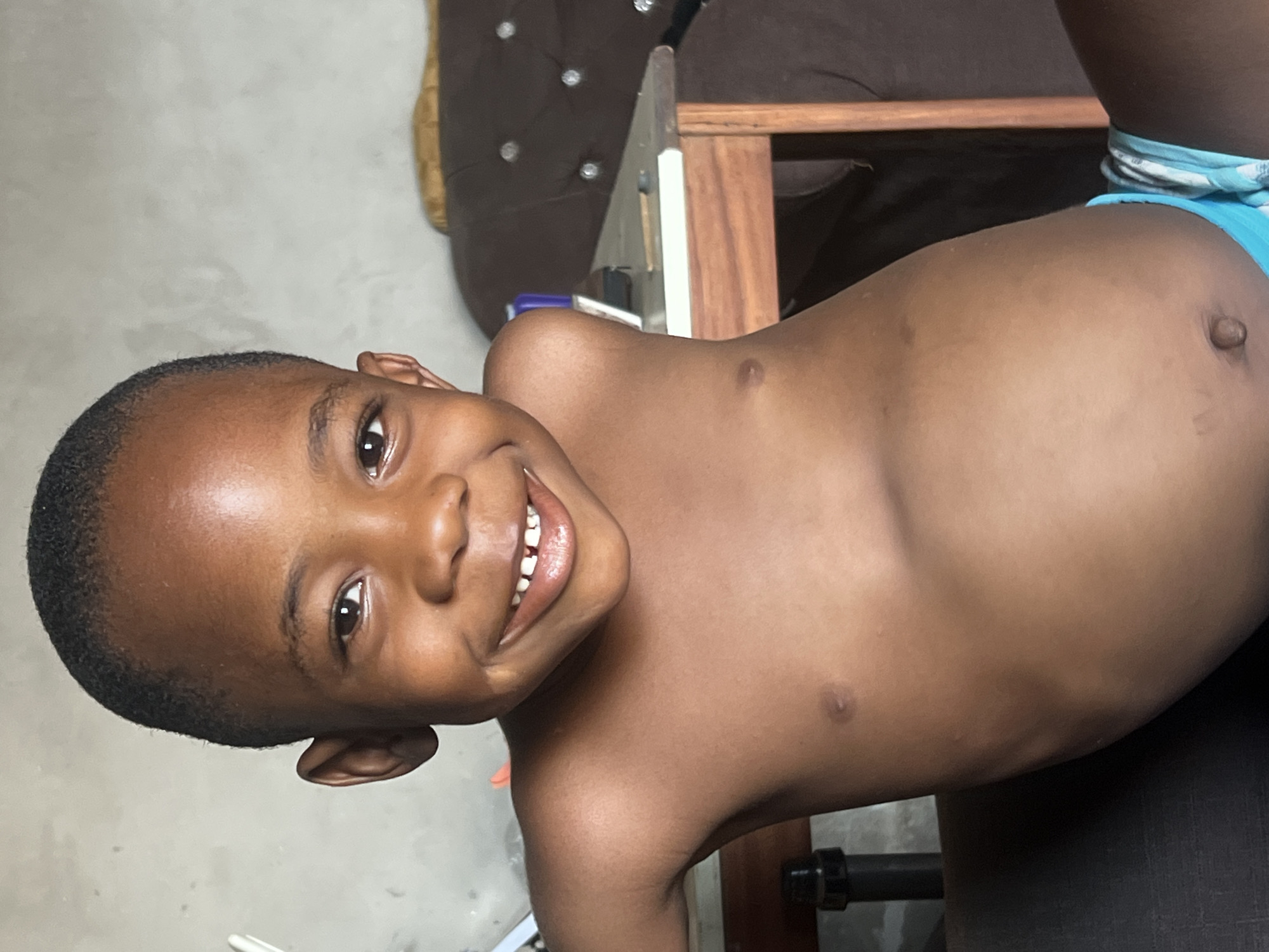 A smiling, young, Black child is pictured from the chest up, facing the viewer, and tilted at an angle. The child has short, dark hair and is wearing blue briefs. The child is situated indoors. Behind the child is a dark, upholstered chair with buttons and a wooden table. A short length of black pipe is partially visible near the lower edge of the frame. The location is described as Minna, Nigeria.