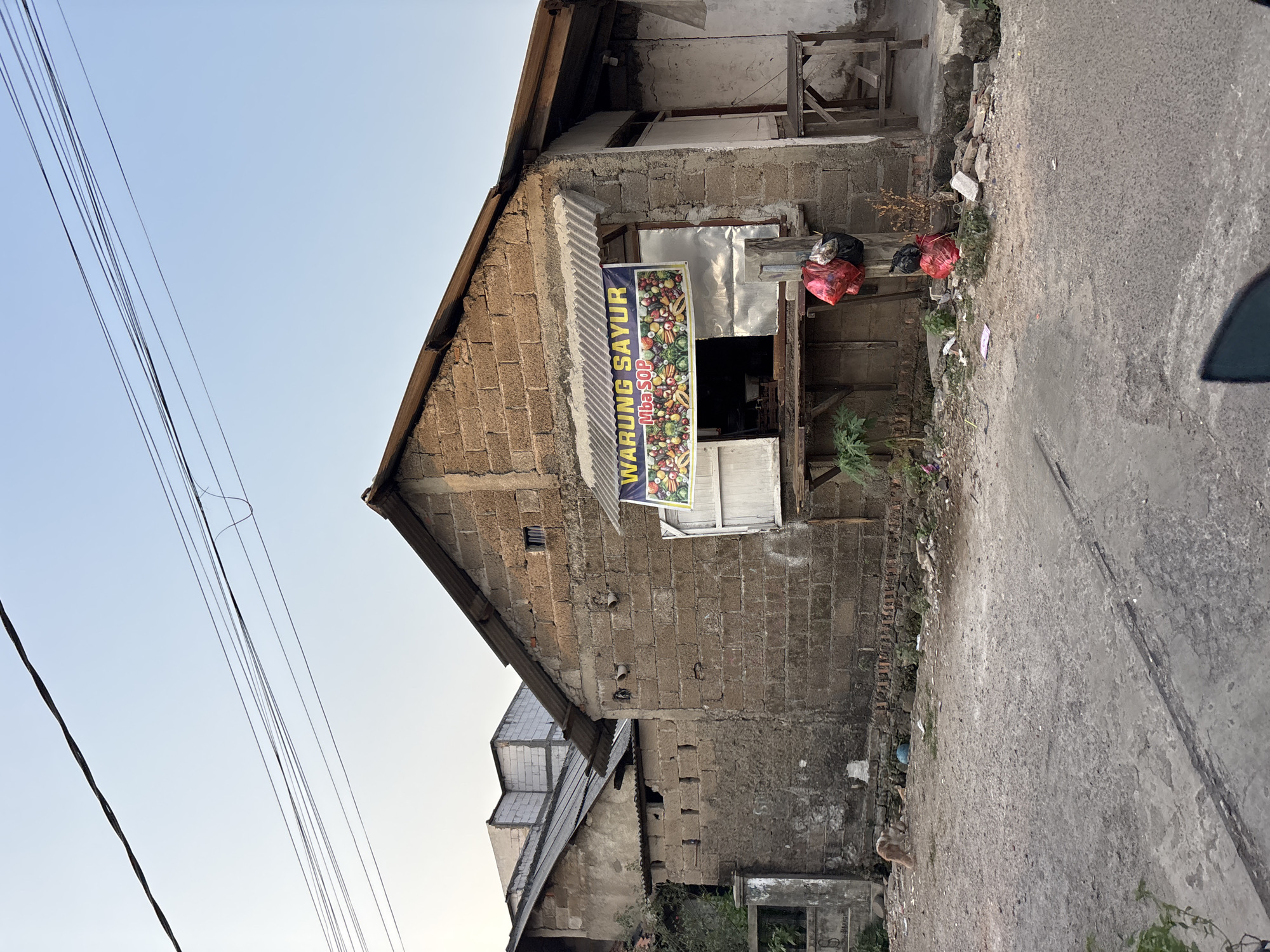 The image depicts a street scene in Bekasi, Indonesia, featuring a small building with a corrugated metal roof that houses a "Warung Sayur" (vegetable stall). A banner above the stall reads "WARUNG SAYUR Mba SOP" and displays an illustration of various vegetables. The building is constructed from rough concrete blocks. In front of the stall, a wooden shelf holds two plastic bags filled with red and black items, possibly garbage. To the left of the stall, a brown and white cat is visible walking away from the camera on a gravelly area littered with debris. Adjacent to the stall, another building with a similar construction style and a darker, sloping roof is partially visible. Utility wires are strung across the sky above the structures. The ground in the foreground is paved with concrete and shows signs of wear and tear, with some cracks and debris. The overall environment appears to be a modest urban setting, characteristic of many neighborhoods in Bekasi.