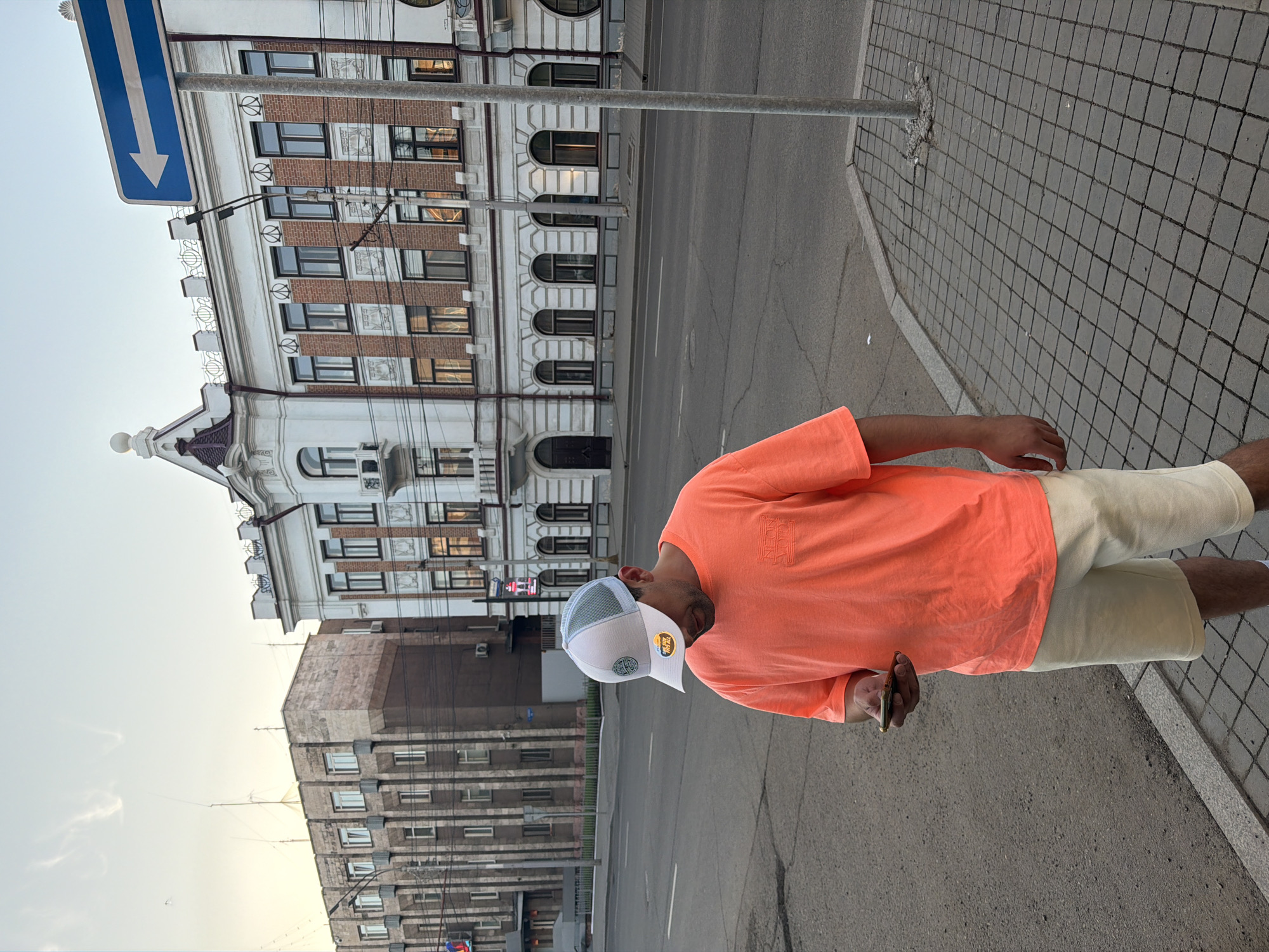 The image, taken in Krasnoyarsk, Russia, captures a daytime urban street scene, focusing on a person on a sidewalk with buildings and a road in the background.
A person, likely male, stands on the right side of the frame, facing slightly left. They wear a bright, coral-orange short-sleeved t-shirt, which has some faded white text on the chest, possibly reading "NEON". They are dressed in light beige or cream-colored shorts. A white and grey mesh trucker-style cap covers their head, featuring a circular logo on the front and another small, yellow-rimmed emblem on the right side of the brim, possibly with text like "THE TEAM". The person holds a dark-colored smartphone in their right hand, looking down towards it or the ground.
The setting includes a patterned grey sidewalk with square paving stones, and a grey asphalt road with white painted lines stretching into the distance. In the background, there are two distinct buildings. Directly behind the person and to the left stands an ornate, multi-story building with a white facade, decorative moldings, arched windows on the lower levels, and brick sections between windows on the upper floors. A blue road sign with a white downward-pointing arrow is prominently displayed on a metal pole in the foreground, partially obscuring this building. Further down the street, to the left, another multi-story building with a more utilitarian, blocky design and numerous windows is visible. The sky above is clear and light, suggesting daytime, possibly late afternoon or early evening. The scene is calm, depicting an everyday city environment.