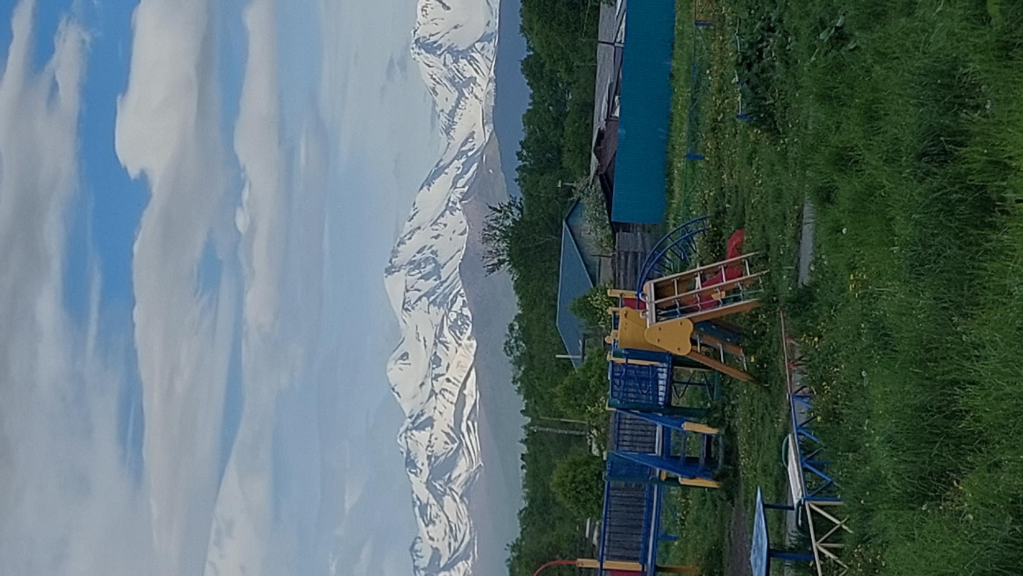 The image depicts a scenic view with a playground in the foreground and snow-capped mountains in the background. The playground features various structures, including a blue and yellow slide, a swing set, and a ladder. Surrounding the playground is lush green grass dotted with yellow wildflowers. In the middle ground, there are several buildings, some with blue roofs, and a turquoise fence. The background is dominated by a majestic mountain range with snow-covered peaks, possibly under a partly cloudy sky. The setting appears to be a rural or semi-rural area, likely in or around Зелёный, Russia, given the presence of the playground and the architectural style of the buildings. There are no people visible in the frame. The overall atmosphere is peaceful and serene.