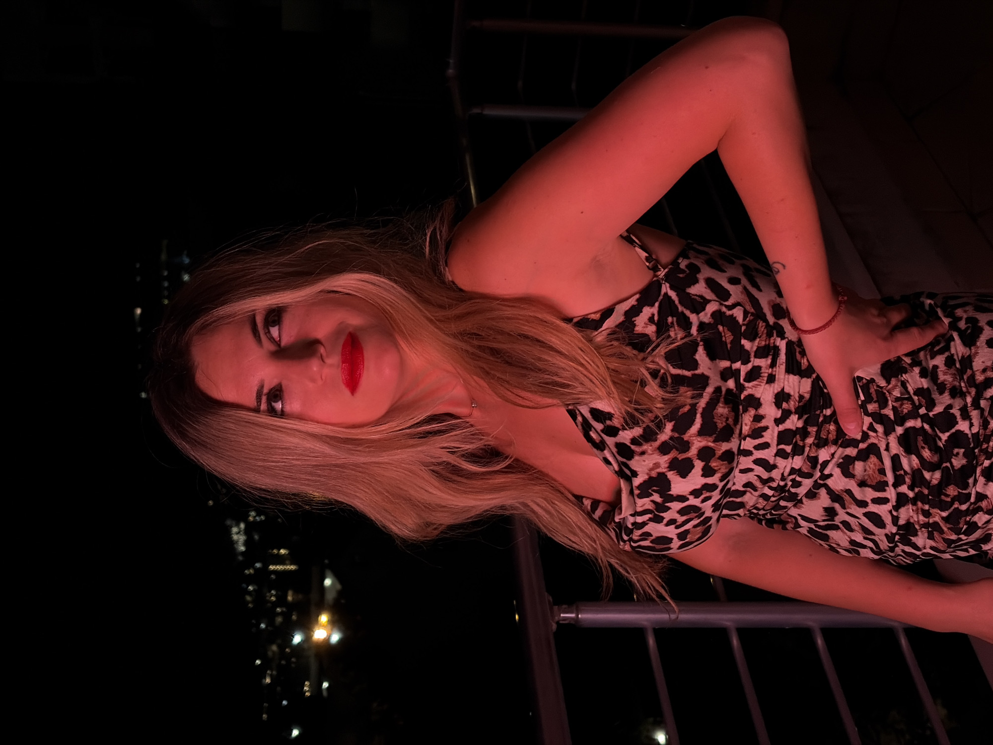 A woman with blonde hair, wearing a leopard print dress and red lipstick, stands on a balcony at night. She is illuminated by red light and looking away from the camera. In the background, the lights of Budva, Montenegro are visible. A metal railing runs along the balcony. Her left hand is resting on her hip, and her right arm is bent at the elbow, with her hand resting on her lower back.