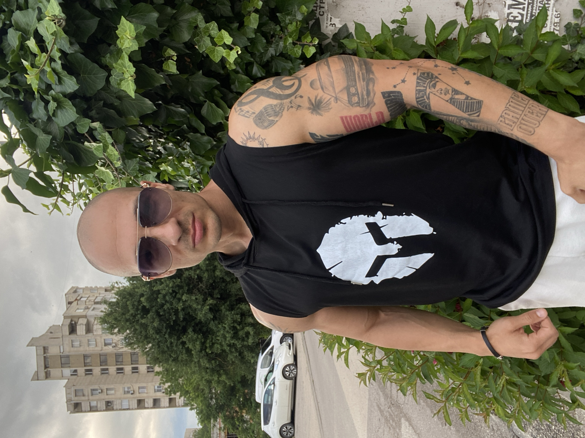 A portrait-style outdoor shot captures a bald man with dark sunglasses posing, looking directly at the camera. He wears a black sleeveless hoodie featuring a prominent white, stylized logo on the chest, and white pants are partially visible. Both of his arms are extensively covered in tattoos, including a microphone, a car, an Egyptian-style bust with star patterns, and text such as "WORLD" on his left arm and "GAME OVER" and "QUEEN" on his right arm. A black bracelet adorns his left wrist.
The man stands next to a wall thickly covered in green ivy. In the background, a street with two white parked cars is visible. Beyond the street, a large, multi-story concrete building, appearing somewhat aged, stands under an overcast sky. Numerous trees and bushes add to the greenery of the urban setting.
The scene takes place during the daytime, likely under cloudy conditions due to the soft, diffused lighting. Visible Cyrillic text on a torn poster or graffiti on the wall behind him includes fragments like "ТЕ И НЕРАЗДЕЛЕНИ В", "Плевен, П", and a number "79.6", confirming the location as Pleven, Bulgaria. The man's expression is neutral and confident.