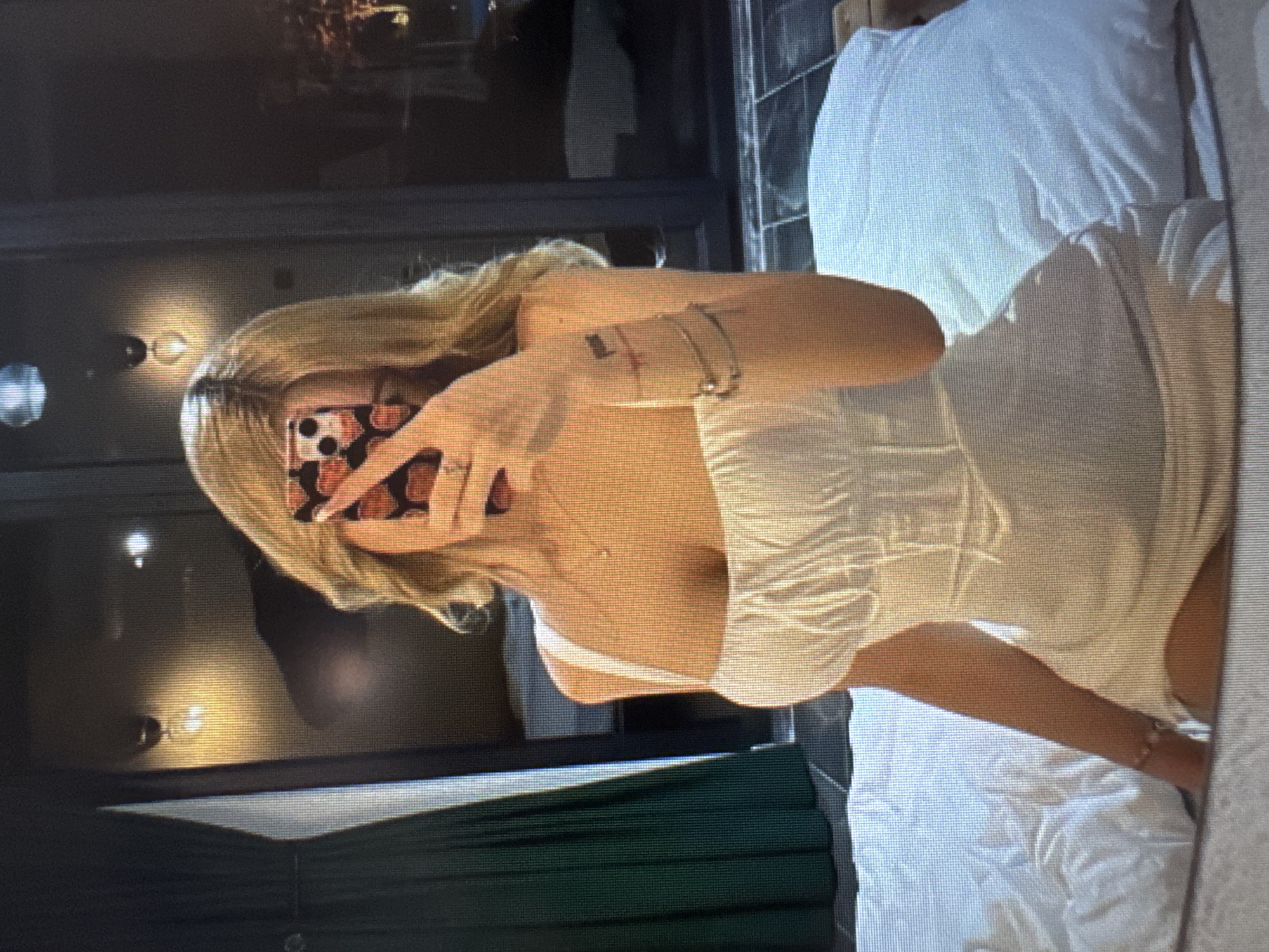 The image, rotated 90 degrees counter-clockwise, depicts a young woman taking a mirror selfie indoors. She has long, blonde hair and is dressed in a light-colored (likely white or cream) strapless or spaghetti-strap top or dress with a ruched bodice. Her face is entirely obscured by the smartphone she holds up to take the photo. The phone is encased in a distinctive black case featuring orange and red floral or abstract patterns and a circular dark element, possibly a PopSocket or part of the camera module.
The woman wears a delicate necklace with a small pendant and a bracelet on her right wrist. A small, dark mark or tattoo is visible on her inner right arm.
The background indicates an indoor setting, likely a bedroom or hotel room. To the right of the woman, a neatly made white bed with white pillows and sheets is visible. The surrounding walls are dark, reflecting warm light from a visible double wall sconce and other unseen fixtures. A dark green fabric, possibly part of a curtain or bedspread, is also present below her. The ambient lighting is artificial and soft, suggesting it is evening or night. The image exhibits a pixelated quality, consistent with a screen capture. No visible text is present.
