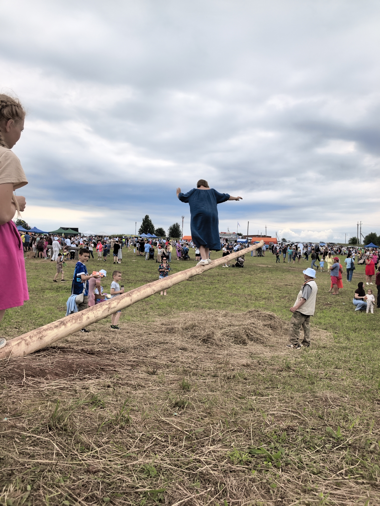 In Кояново, Russia, during an outdoor event, a woman in a blue dress is balancing on a long, thick log positioned across a grassy field. The log is angled, with its higher end supporting the woman. In the foreground, a young girl with blonde braided hair stands on the left, facing away from the camera and towards the event. Several children and adults are scattered throughout the background, with tents and structures visible in the distance, suggesting a festival or fair. The sky is overcast with dramatic clouds. The ground is covered in dry grass and some scattered hay.