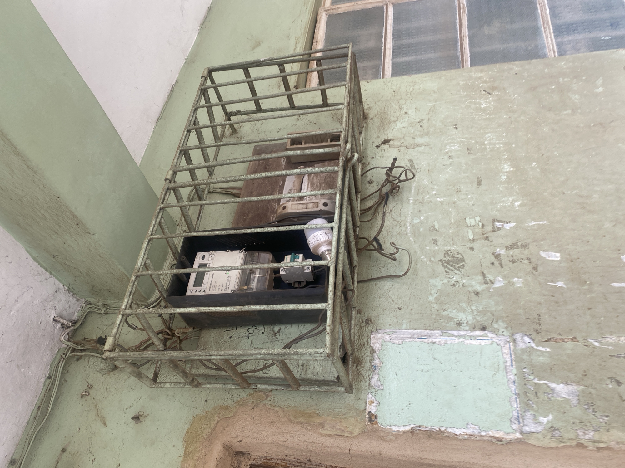An exterior shot shows a weathered, pale green painted wall in Abeokuta, Nigeria. The wall has areas of peeling paint, exposing a lighter colored surface beneath. To the right, a window with multiple panes of frosted glass is visible, set within a similarly colored frame. Prominently featured is a rusty, metal cage mounted on the wall, enclosing electrical equipment. Inside the cage, a white electricity meter with digital display and some wiring is visible. To the right of the meter, a wooden box or panel is also present within the cage. Loose electrical wires hang from the cage and connect to conduits running along the ceiling and down the wall. The overall impression is one of disrepair and utilitarian design.