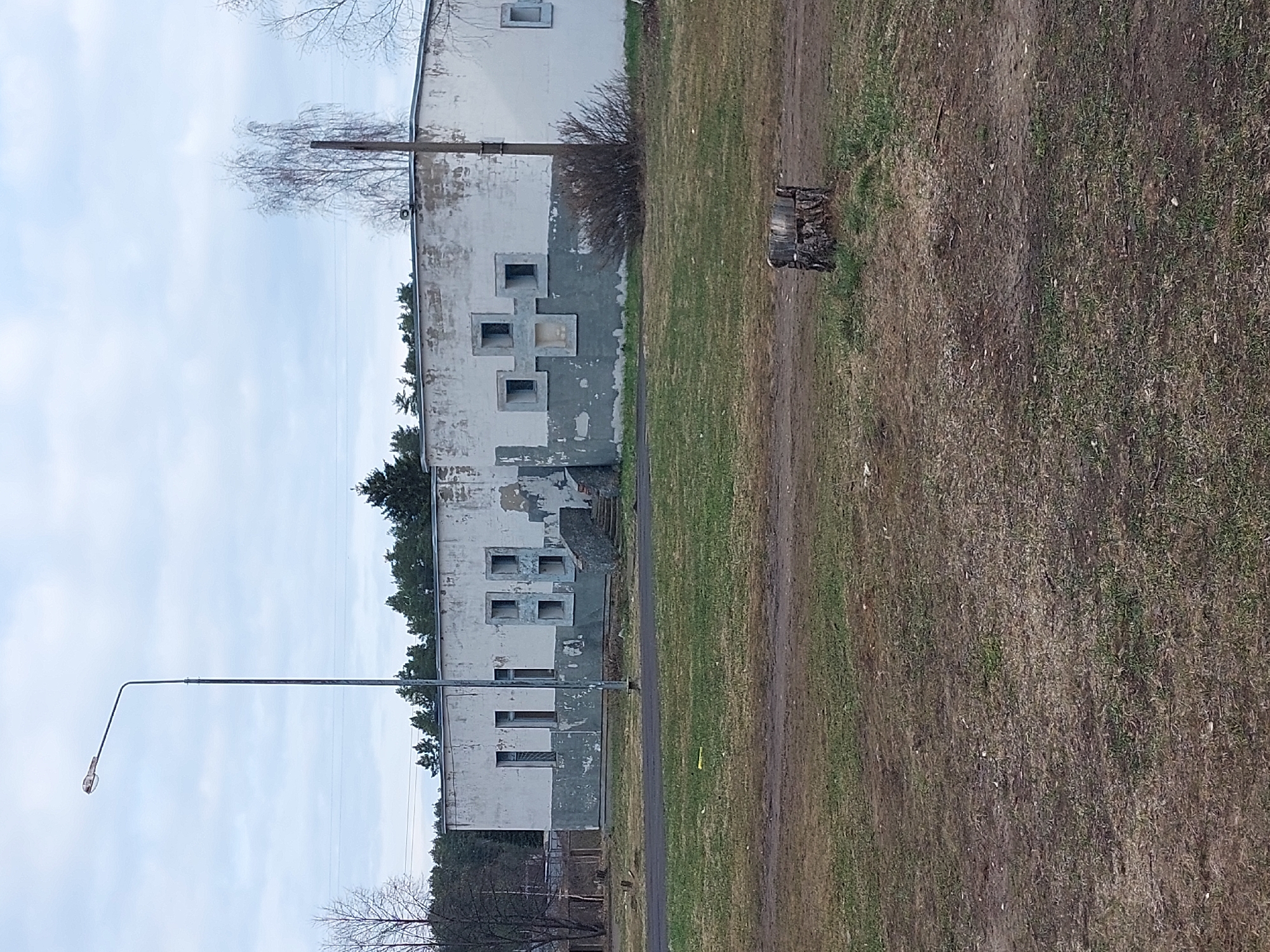 The image shows a multi-story building with faded white and gray paint in Chaikovsky, Russia. There are rectangular windows, some with concrete frames. A dirt path runs alongside the building. Grass grows along the path. A tree stump is visible on the path. A street light stands to the left of the building. The sky is overcast with thin clouds. Trees are visible in the background.