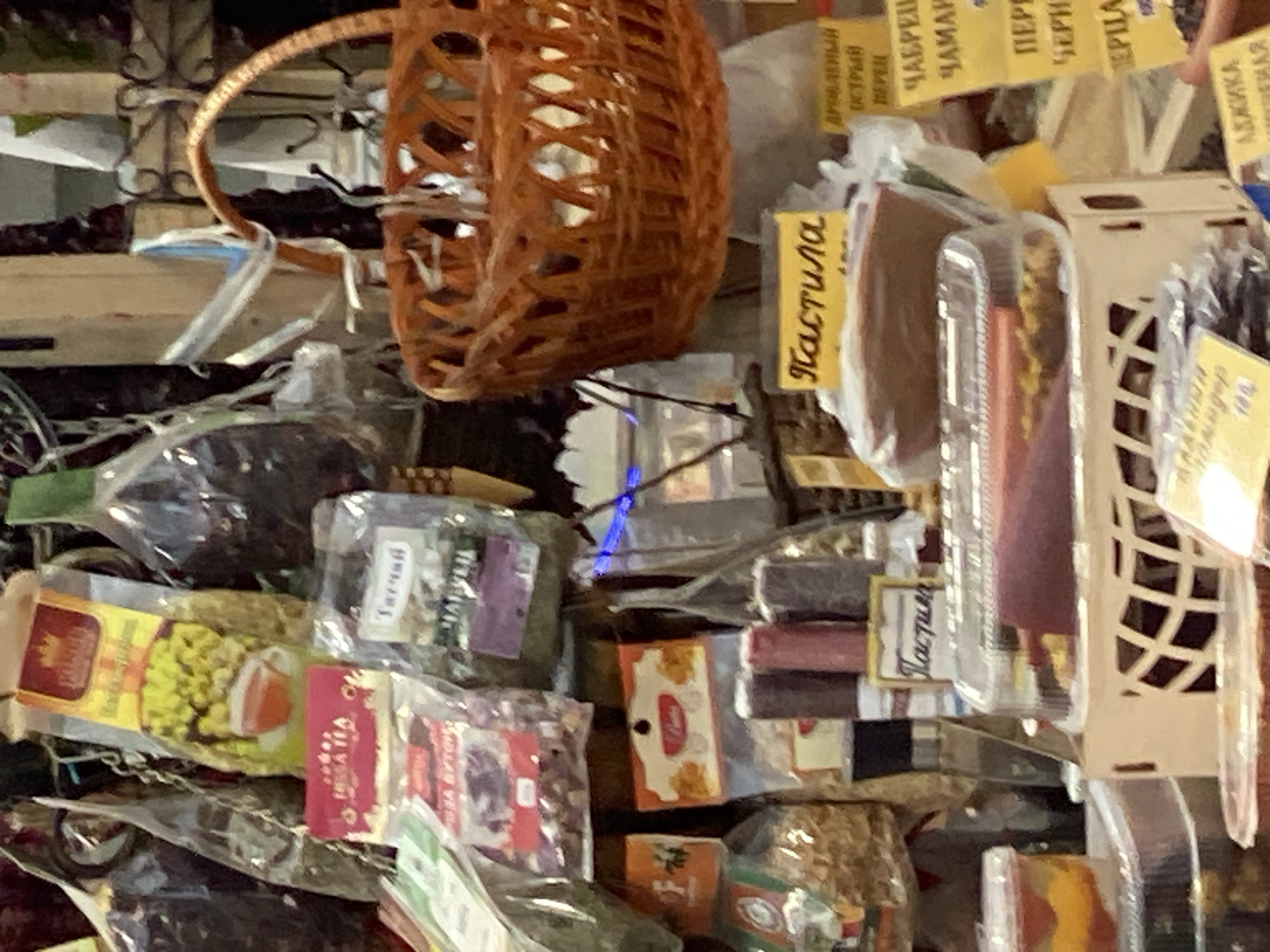 This image depicts a close-up view of a stall filled with various packaged goods, likely herbs, spices, or dried fruits, arranged for sale. A woven basket is prominently featured at the top, with a rustic texture. Below the basket, numerous transparent plastic packages contain the items, some with visible labels in Cyrillic script. The overall impression is of a market or small shop specializing in traditional or artisanal products.
Several labels with text are visible. One prominent label in yellow reads "Пастила," which translates to "pastila," a fruit confection. Other visible text includes "ЧАБРЕЦ" (chamomile), "ЧАМАР" (a type of thyme), "ПЕРЦА" (pepper), "АДЖИКА" (adjika, a spicy paste), and "Тагчий" which may refer to a specific herb or spice. The packaging suggests a focus on natural or homemade items. The lighting appears to be natural, possibly indicating an indoor setting or an overcast day, as there are no strong shadows. The context provided mentions Novaya Usman', Russia, which aligns with the Cyrillic script. The image captures a detailed snapshot of the products offered at this particular vendor.
