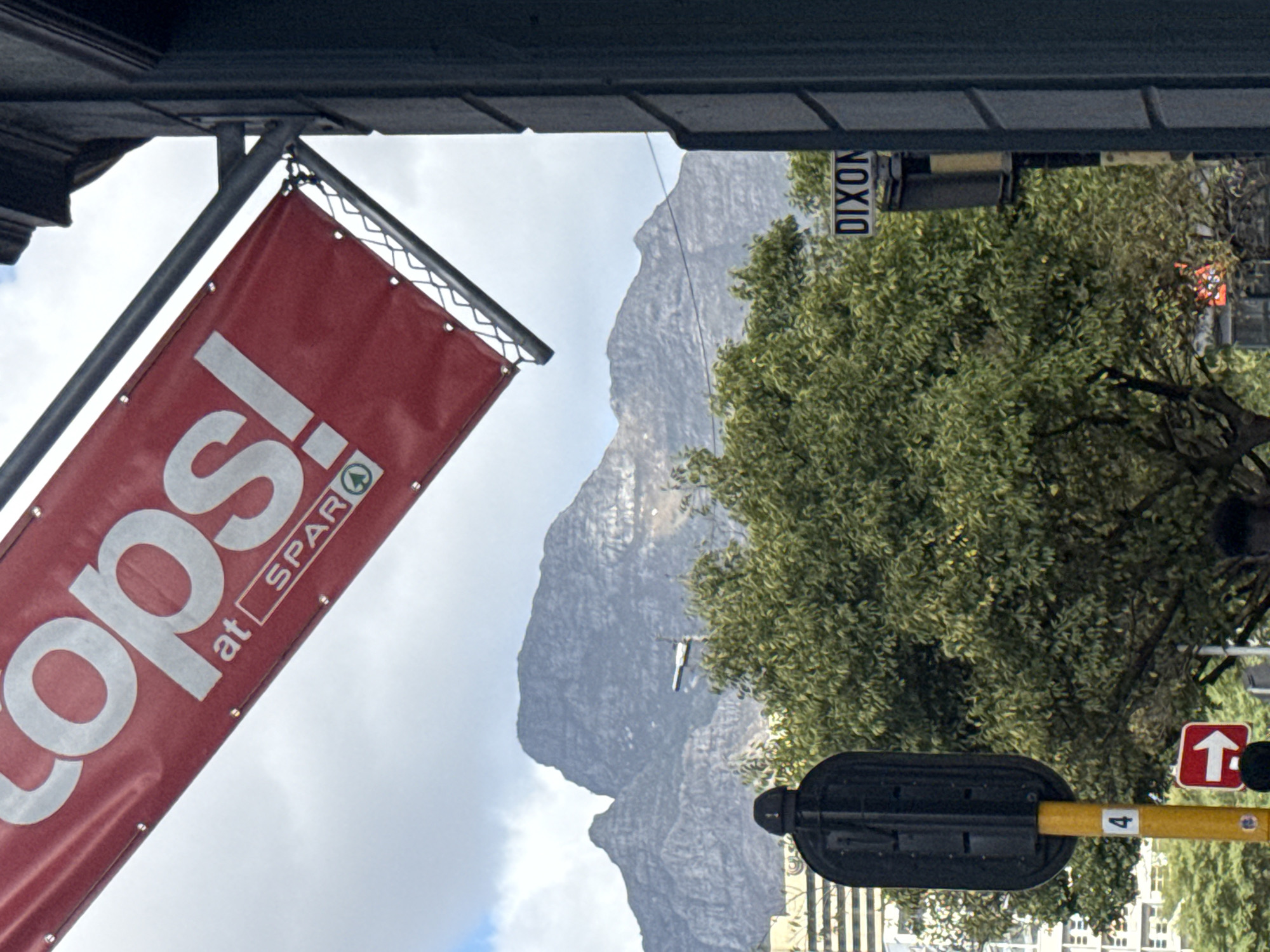 A slightly angled view of a scene in Cape Town, South Africa, captured beneath an overhang. A large red banner with "TOPS! at SPAR" displayed in white, hangs on the left. The background features Table Mountain under a cloudy sky. A street sign reading "DIXON" is visible, partially obscured by a green tree. A black traffic signal fixture with a yellow pole, marked with a "4" and an upward-pointing arrow, is also present. In the far background, buildings appear at the base of the mountain.