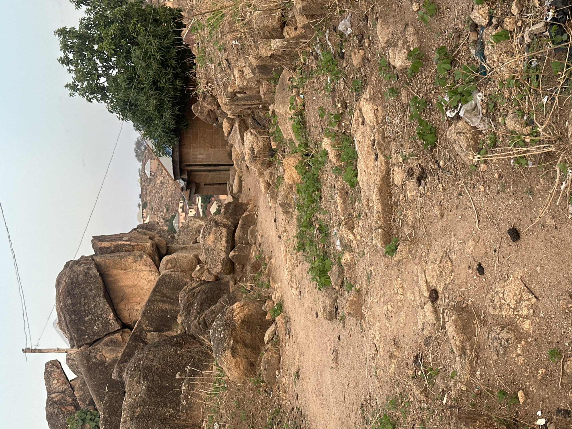 This is an outdoor shot of a rocky, sandy path in Jos, Nigeria. The path leads uphill, flanked by large boulders and sparse vegetation. A small, brick building is visible on the left, nestled among the rocks. In the background, a cluster of buildings suggests a settled area on higher ground. Litter, including plastic debris, is scattered along the path. Dark objects, possibly animal droppings, are scattered on the ground. A utility pole with wires is visible on the left. The sky is overcast, suggesting a hazy or humid atmosphere. No people are visible in the frame.