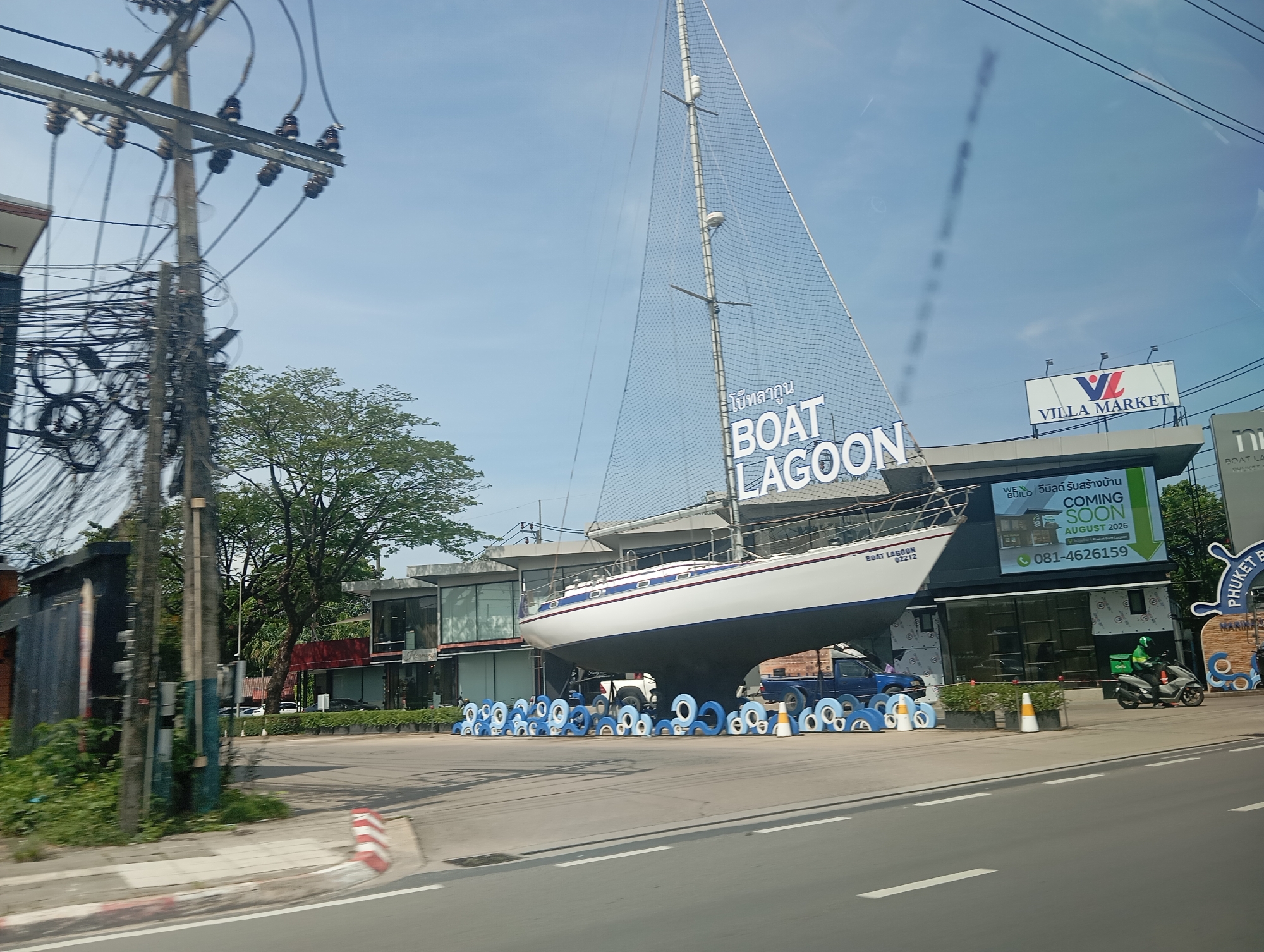 A sailboat is mounted on a structure in front of a commercial building in Ko Kaeo, Thailand. The sailboat is white with a blue stripe and has the words "BOAT LAGOON 02212" on its hull. In front of the building is a large sign that reads "VILLA MARKET COMING SOON AUGUST 2026 081-4626159". A person on a scooter with a green bag is riding on the road in front of the building. The sky is blue and clear. There are utility poles and wires in the foreground.