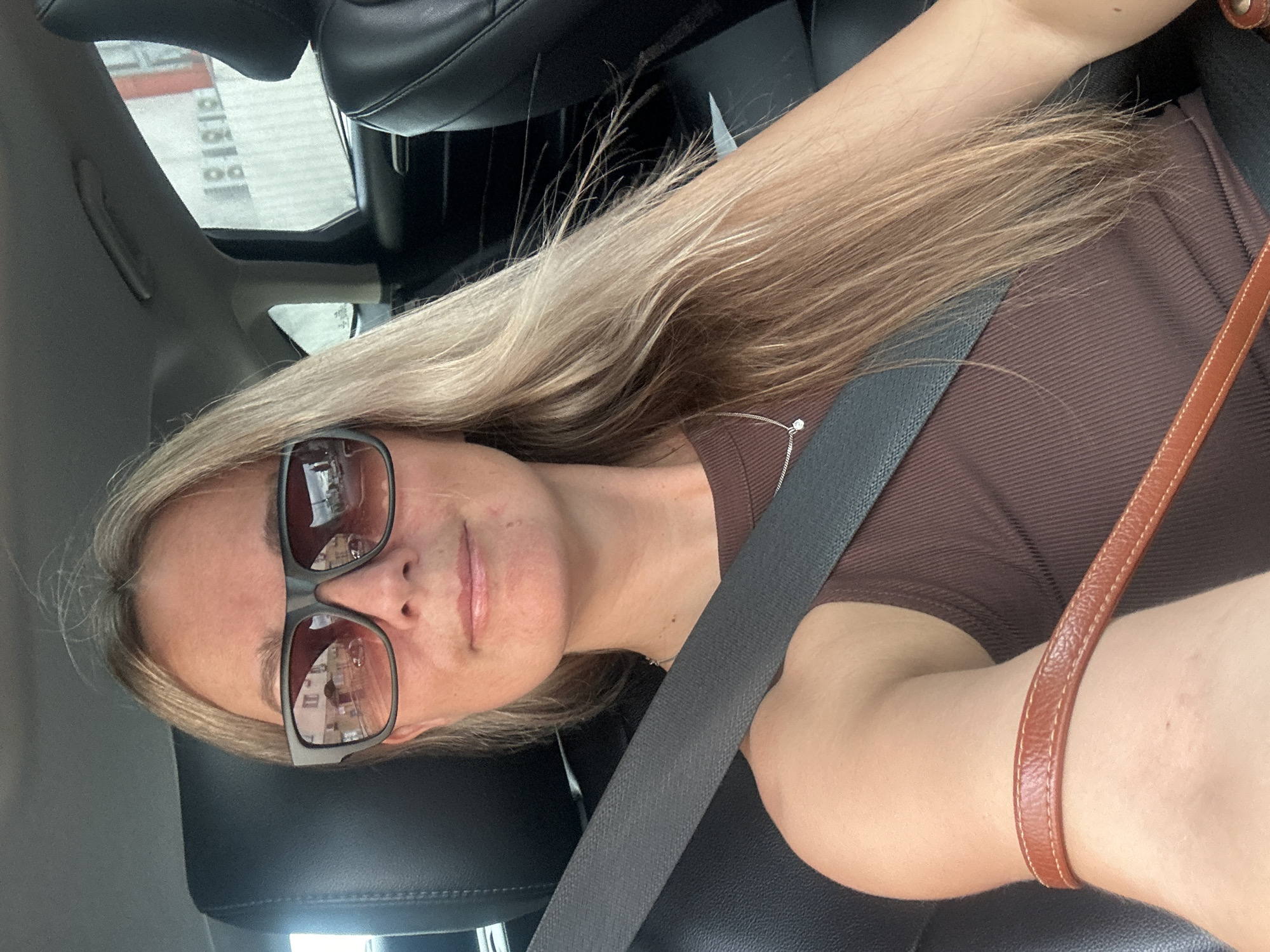 A woman with blonde hair and sunglasses is sitting in the passenger seat of a car. She is wearing a brown tank top and a silver necklace. A seatbelt is fastened across her chest. A brown purse strap is visible on her shoulder. In the background, through the car window, buildings and street signs are visible, suggesting an urban environment. The image does not provide any specific visual cues to pinpoint the location as Sochi, Russia.