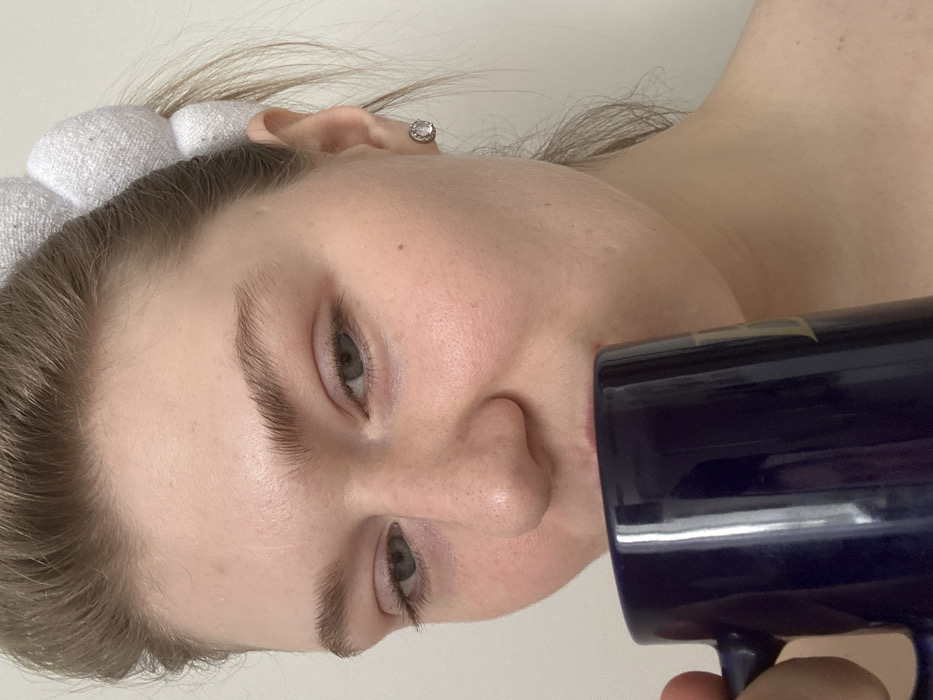 The image captures a close-up, rotated portrait of a person, likely female, appearing to drink from a dark blue mug. The person has fair skin, light brown hair, and neutral facial features with open eyes. Their hair is partially pulled back by a white, soft-textured headband, and a single stud earring is visible in their right ear. The dark blue mug is held to the person's mouth, with its handle visible towards the bottom right of the frame. A subtle lighter-colored band or tape is visible on the side of the mug. The background is a plain, light color, indicating an indoor setting. Only the upper chest and shoulder area are visible, appearing bare, suggesting casual attire. The overall expression is relaxed. No visible text is present in the image. The location context provided is Kazan, Russia.