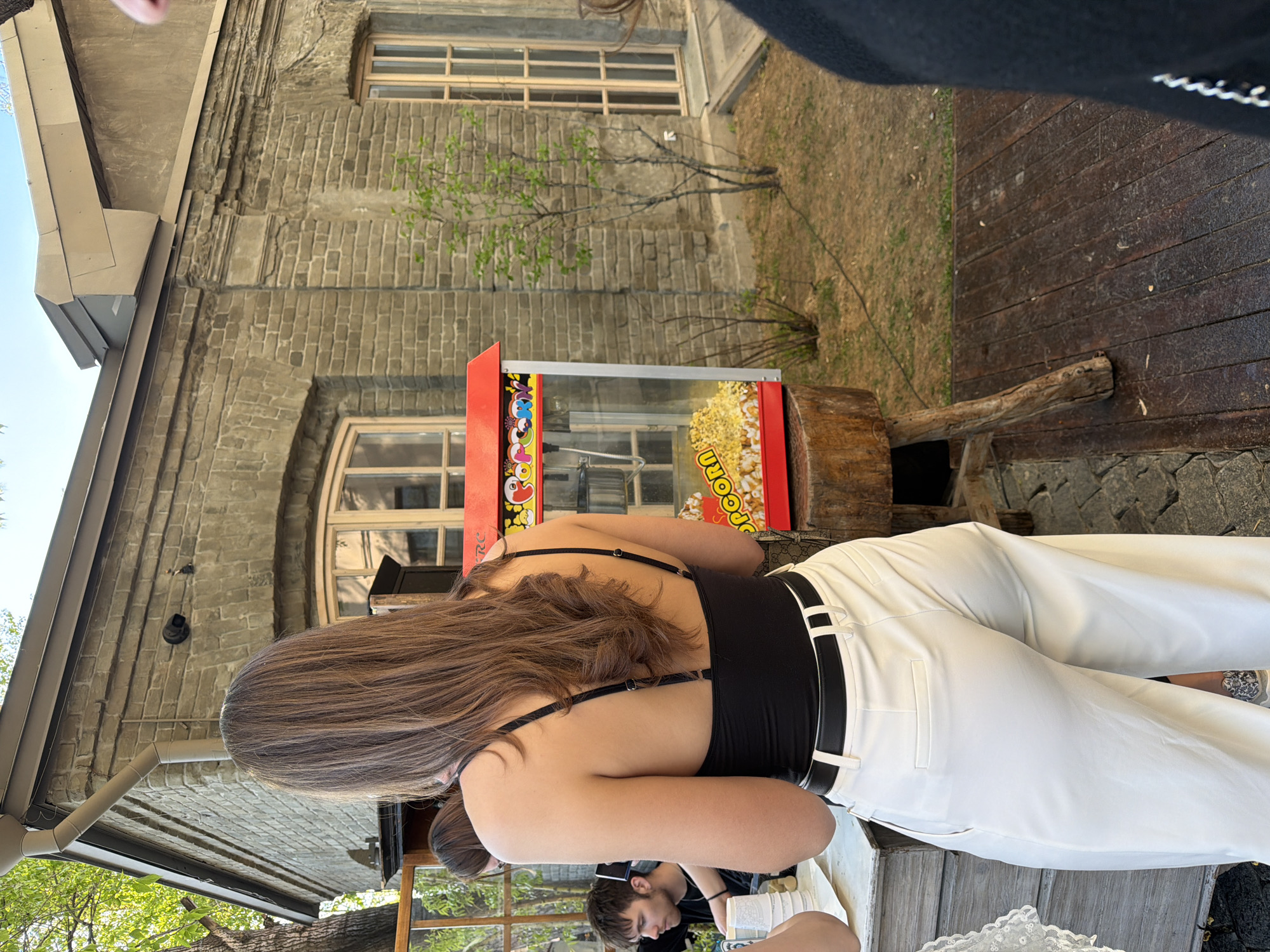 A slightly tilted, eye-level outdoor shot depicts a young woman with long brown hair, wearing a black top and white pants with a black belt, standing near a popcorn machine outdoors. The popcorn machine is next to a tree stump. The building behind is made of tan brick, and has a large arched window. Other people are partially visible at the bottom of the image.
Based on the architectural style and the presence of a popcorn machine in an outdoor setting, it is plausible that the location is within Moscow, Russia, possibly at a cafe or public space.