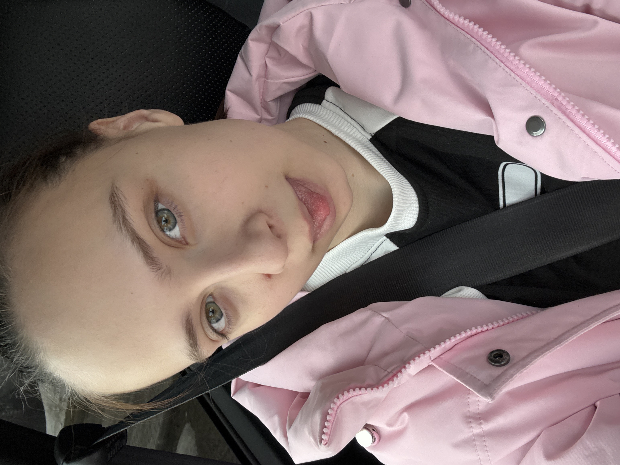 A young person with fair skin, light brown hair, and green eyes is positioned in what appears to be the passenger seat of a car, based on the presence of a black seatbelt across their chest. They are wearing a pink hooded jacket over a black and white top. The background is mostly dark and out of focus. There is no indication of the image being taken in the city of Прикамский, Russia.