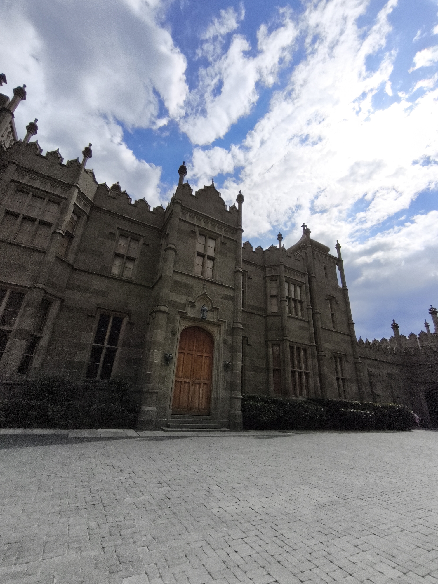 The image depicts the Vorontsov Palace in the city of Алупкинский городской совет, Ukraine. The palace, built in the English Gothic style, features gray stone walls, ornate windows, and decorative battlements. A prominent wooden door is located at the center of the facade. The sky above is partly cloudy, with patches of blue visible. The foreground consists of a paved courtyard made of gray cobblestones. A low hedge of dark green bushes runs along the base of the palace.
