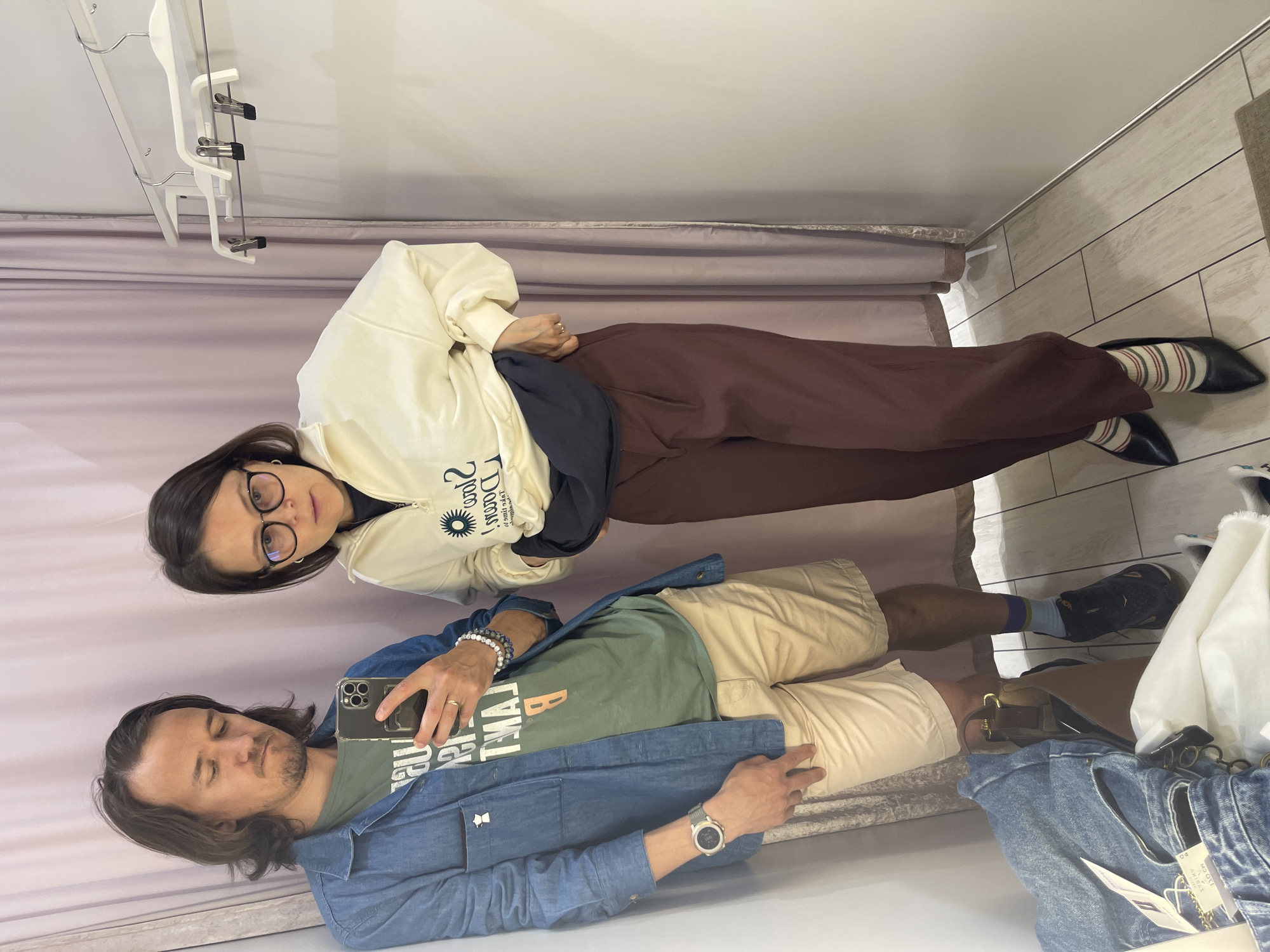 Two people are in a fitting room. A woman with dark hair and glasses is wearing a white sweatshirt, a black shirt, and brown pants, with striped socks and black shoes. A man with long hair is wearing a blue jacket, a green shirt, and tan shorts, and is holding a phone. The fitting room has a purple curtain and a hanger rack.