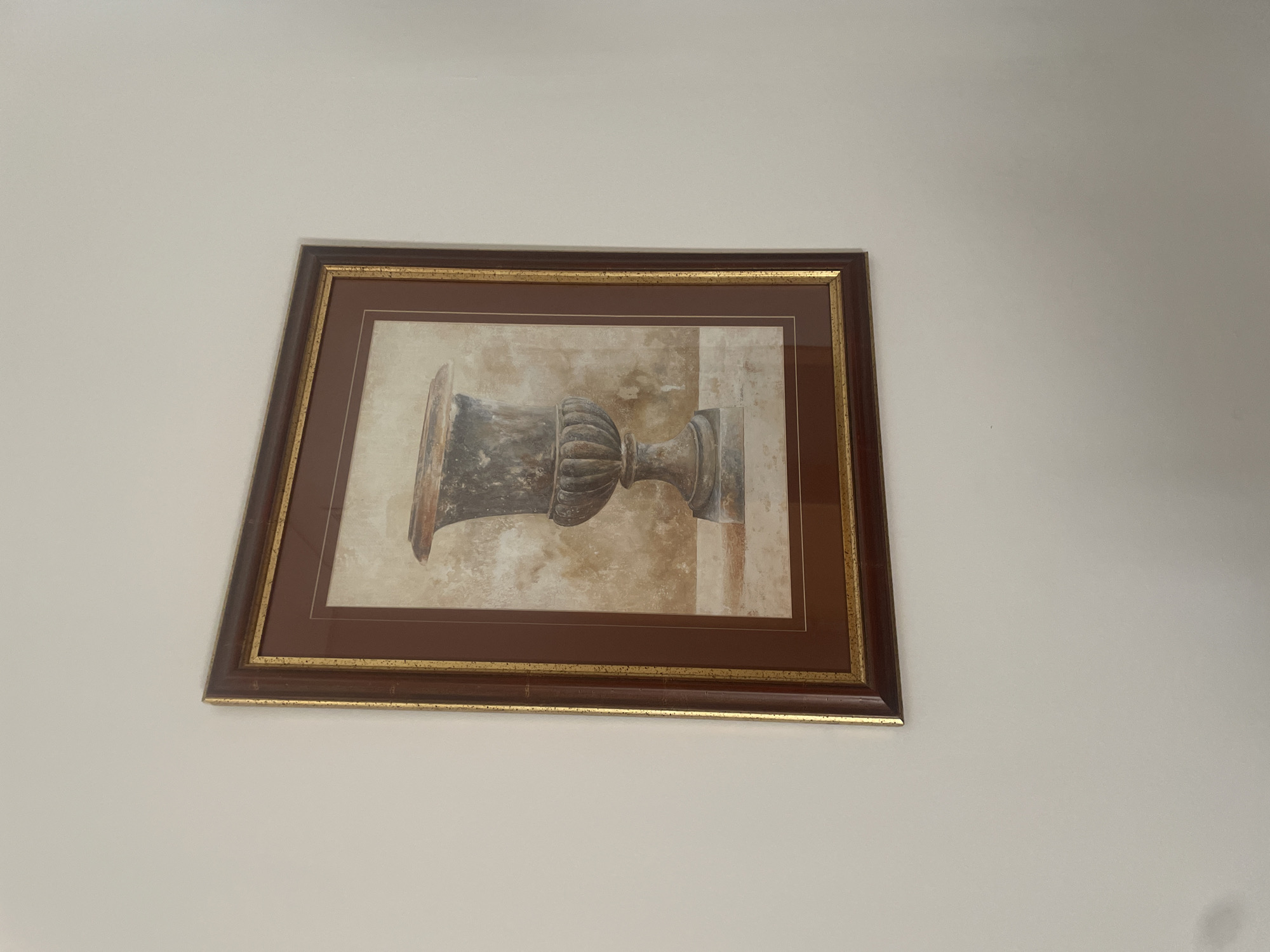 A framed painting of an urn hangs on a plain, light-colored wall. The painting features a dark-colored urn with decorative fluting on its base and a wide opening. The background of the painting is textured and appears to be in muted earth tones. The frame is dark brown with a thin gold inner border. The image is taken from a slight angle, showing the top left corner of the frame. The scene appears to be indoors. There is no information available to indicate this is located in Sevilla, Spain.