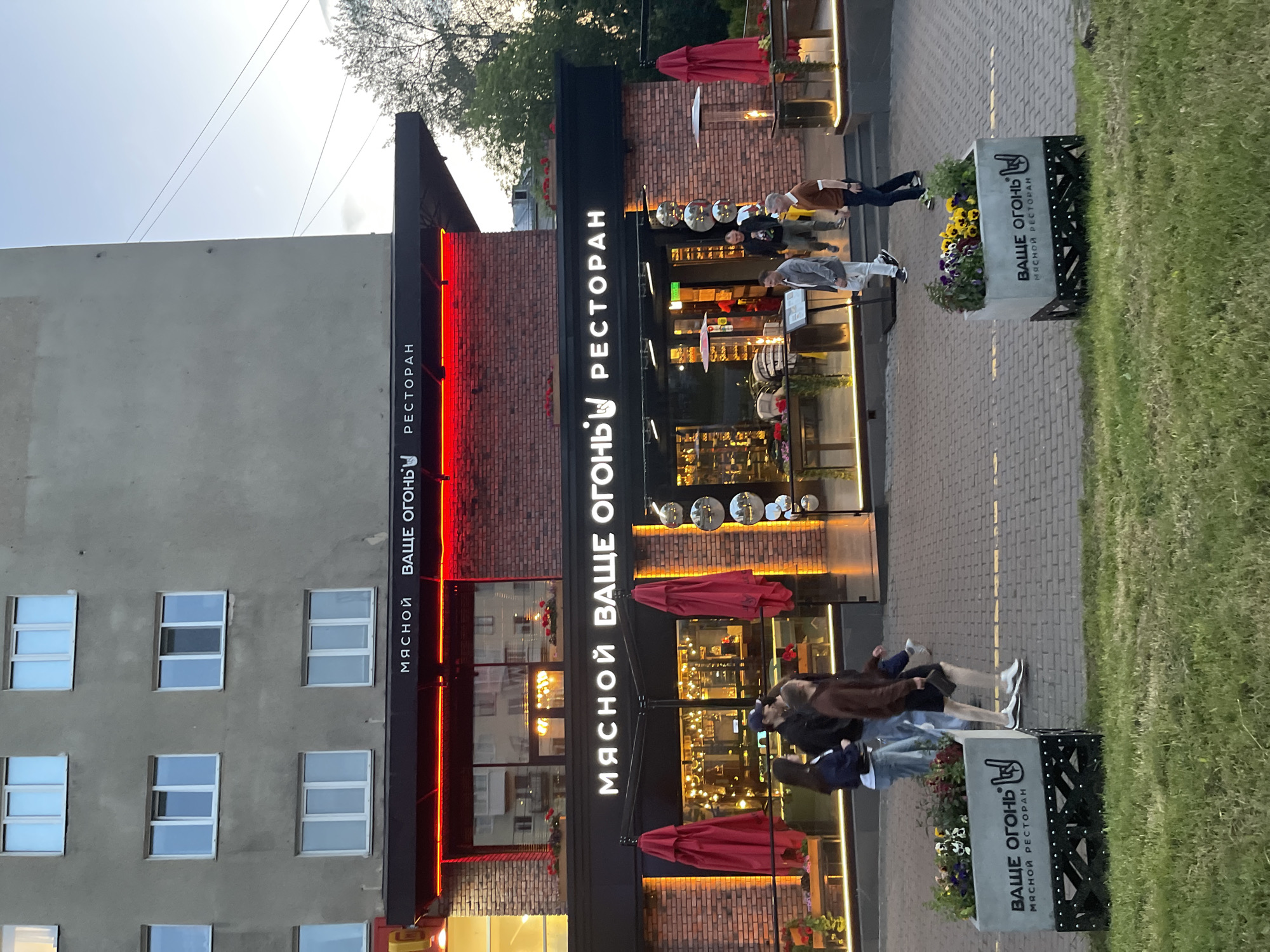 The image shows the exterior of a restaurant called "Вкусный Огонь" (Vkusnyyogon), which translates to "Tasty Fire." The restaurant has a modern design with a brick facade and large windows. Red umbrellas and outdoor heaters are visible on the patio. Several people are walking by the restaurant and entering and exiting. The scene appears to be in a city, with a multi-story building in the background. The lighting suggests it is either early morning or late evening.