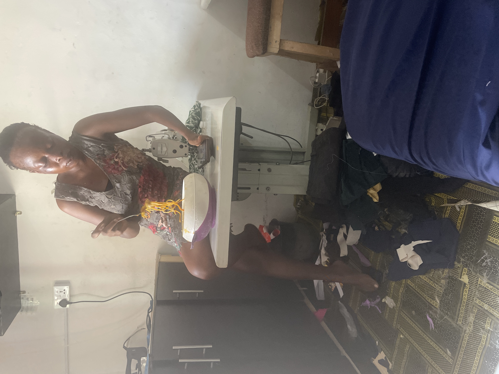 A woman sits on the edge of a sewing machine table, eating noodles from a bowl. She is wearing a floral-patterned dress. The room appears to be a workshop or tailoring space, with fabric scraps and a sewing machine visible. The floor is covered in a patterned rug, with more fabric and debris scattered around. The room is sparsely furnished, with cabinets and a chair. The lighting is somewhat dim, with an electrical outlet visible on the wall.