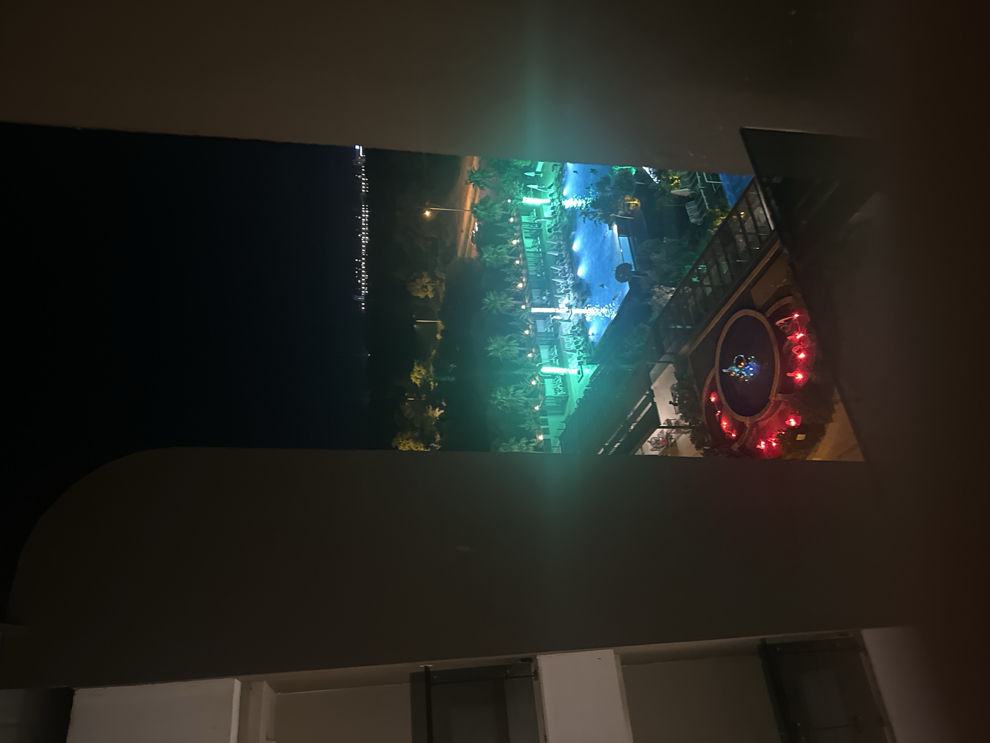 The image depicts an aerial view of a resort at night, possibly in Manavgat, Turkey, as suggested by the prompt's context, though the location is not explicitly identifiable within the image itself. The scene is framed by dark, indistinct architectural elements, suggesting the photograph was taken from a window or balcony.
In the foreground, a circular fountain with red lights and water jets is visible, adding a splash of color against the darkness. Beyond the fountain, a large swimming pool is illuminated with vibrant green lights, creating a striking contrast with the surrounding night. Several lounge chairs with umbrellas are neatly arranged around the pool area. Some individuals are present in the pool, indicating activity despite the late hour.
The background features a line of trees and palm trees, typical of a tropical or subtropical resort setting. Further in the distance, a faint outline of a city skyline with scattered lights suggests proximity to an urban area or another developed settlement. A road with streetlights and a few cars can be observed, hinting at the resort's accessibility. The overall impression is one of a lively resort complex illuminated for nighttime enjoyment, set against a dark, natural, and distant urban landscape.