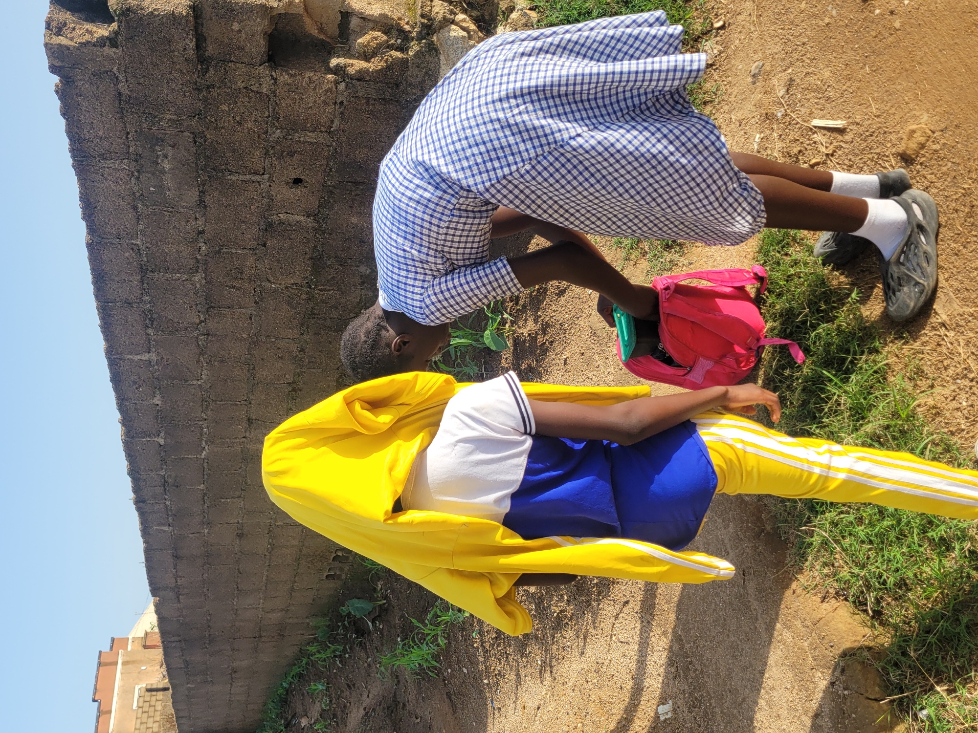 Two young individuals are near a low, rough-hewn stone wall in what appears to be an outdoor urban setting. The person on the left, facing away from the camera, is partially covered by a bright yellow garment draped over their head and shoulders, obscuring their face. They are wearing a blue and white top and yellow pants with white stripes. The person on the right, wearing a blue and white checkered dress and white socks with dark sandals, is bending over and appears to be placing an item into a bright pink backpack. The ground is a mix of dirt and sparse green grass. In the background, a simple brick building is visible beyond the stone wall. The lighting suggests it is daytime, possibly morning or late afternoon. The scene is likely within the city of Jos, Nigeria, given the architectural style and vegetation.