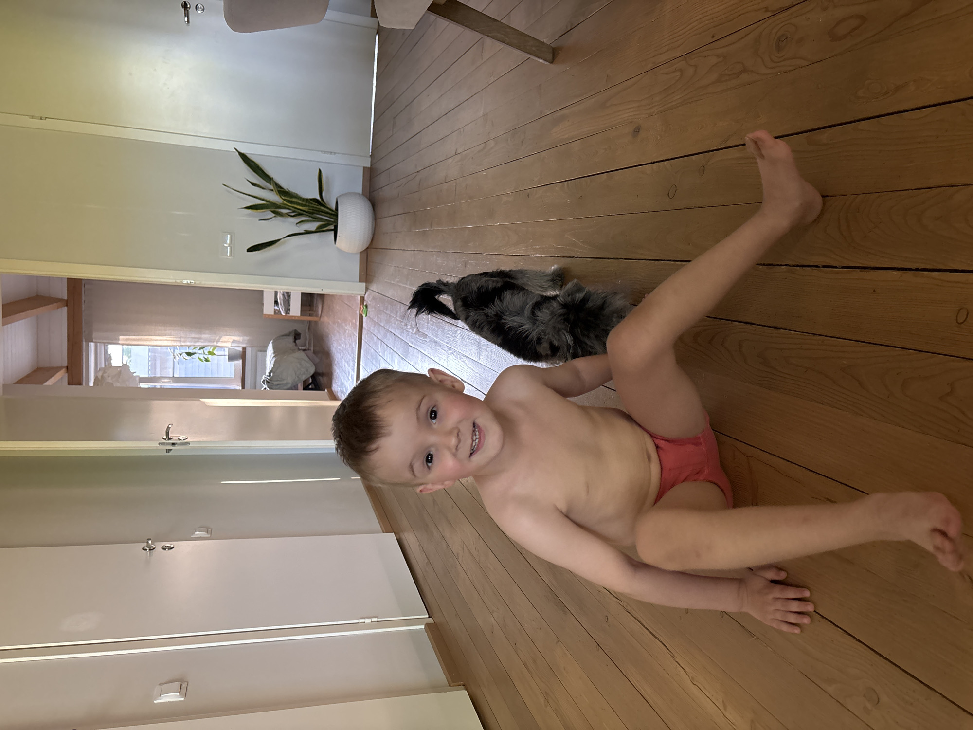A child, presumably male, lies on a wooden floor, clad in what appears to be pink underwear or a diaper. He is smiling and looking towards the camera. A small, dark-colored dog is partially visible behind him. The room appears to be indoors, with a white door and a potted plant visible in the background. The scene is illuminated by natural light. It is impossible to ascertain if this scene is in the city of Chaikovsky, Russia.
