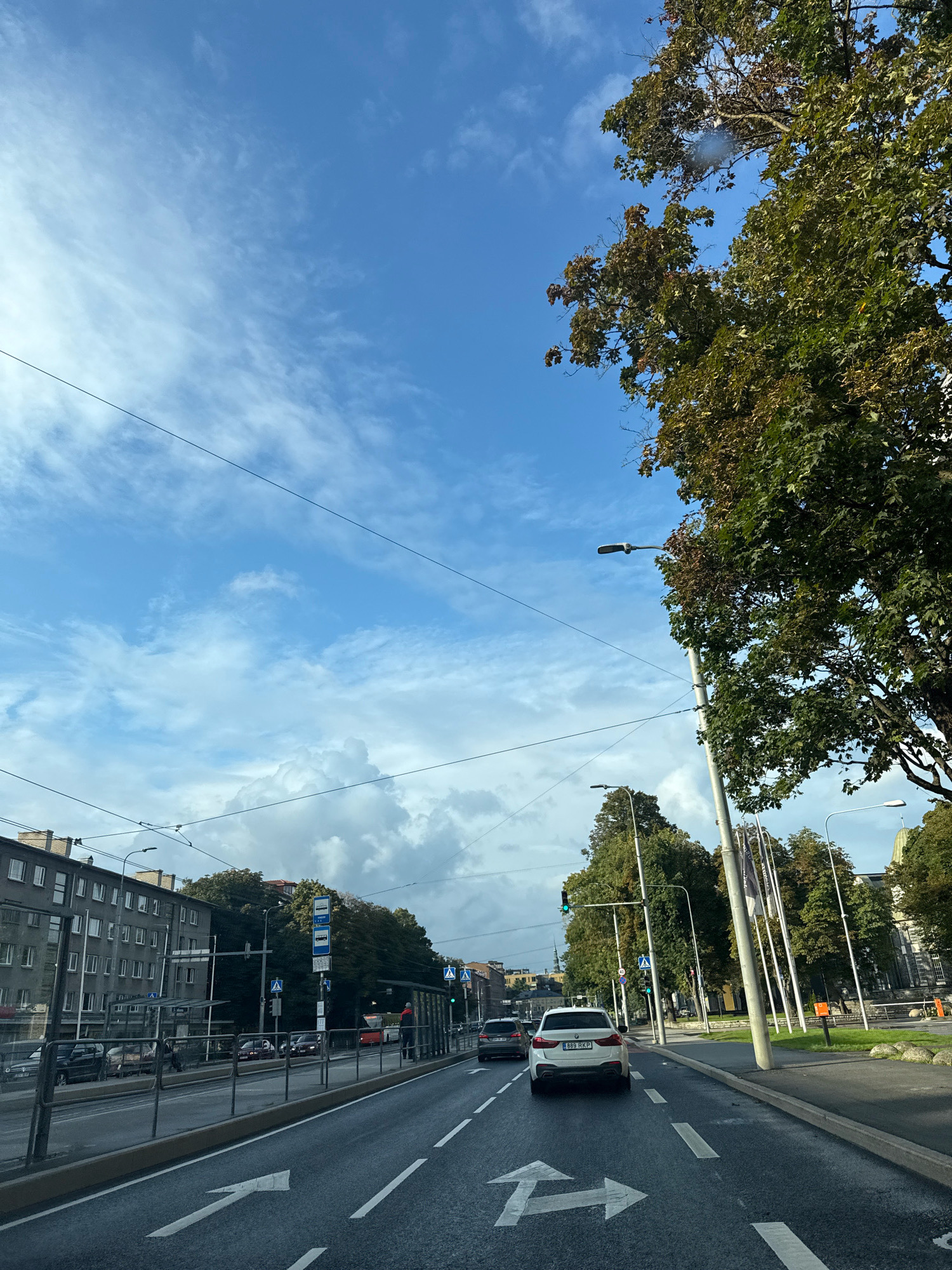 The image depicts a street scene in Tallinn, Estonia, under a partly cloudy sky. The perspective is from within a vehicle traveling on a multi-lane road. In the foreground, the asphalt road features white directional arrows. To the left of the road, there is a sidewalk with a metal railing separating it from the traffic lanes. Behind the railing are parked cars and a modern building with multiple windows. Further down the street, there is a bus stop with a shelter and signage indicating bus routes. Trees with green and brown foliage line the sidewalks and median. To the right of the road, there is a grassy area with some large rocks and several tall poles, possibly flagpoles, and a streetlamp. The sky is a bright blue with scattered white clouds, and some trees with dense green leaves are visible, with their branches extending into the upper portion of the frame. A white SUV is in front of the viewer's vehicle, and other cars are visible in the distance. The overall impression is an urban environment on a clear day, likely during autumn given the tree foliage.