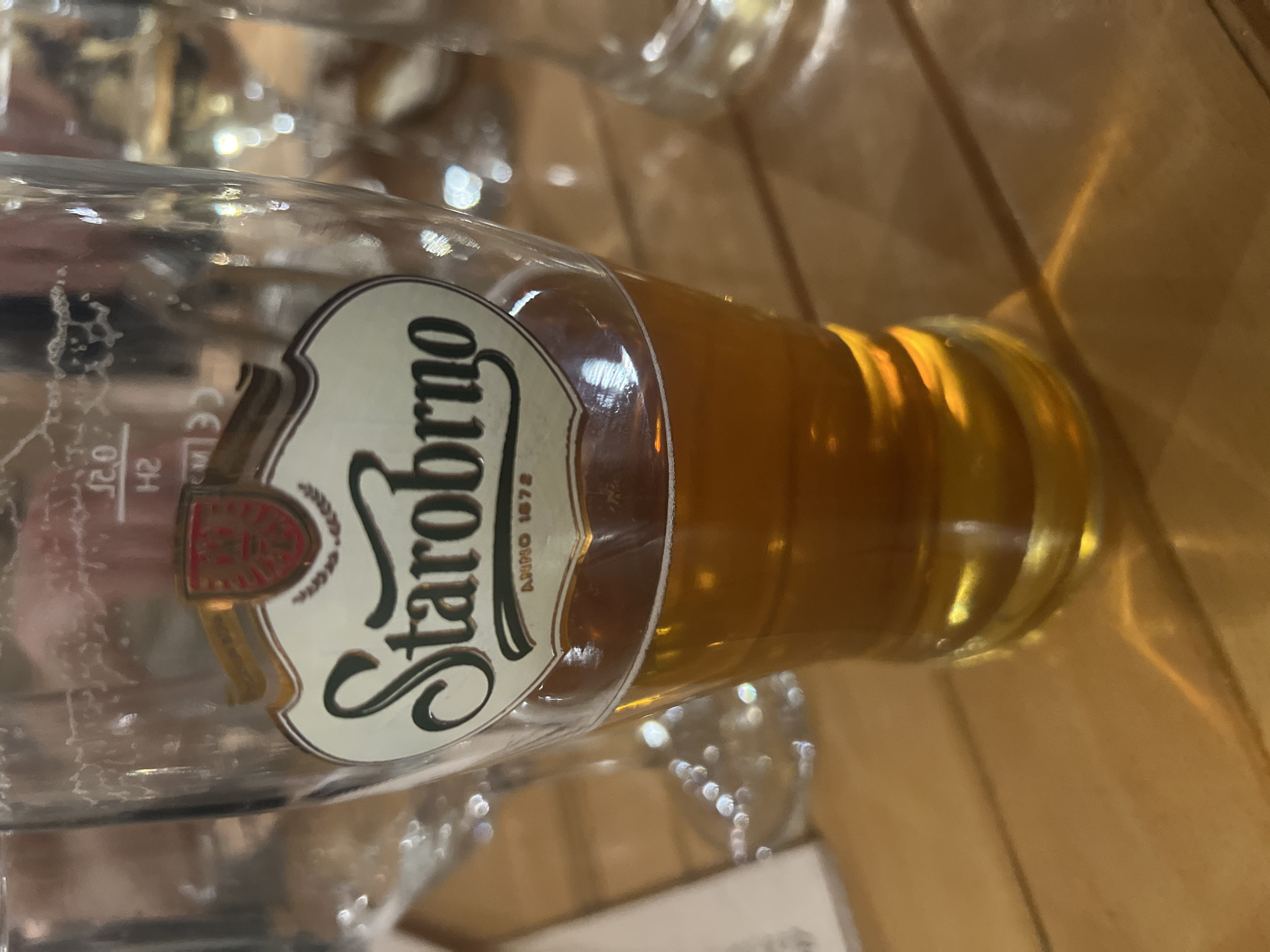 A close-up, tilted shot captures a glass of Starobrno beer, partially consumed, resting on a warm-toned wooden surface in Sofia, Bulgaria. The glass holds a rich amber liquid at its base, reflecting ambient light. The prominent label on the glass features the brand name "Starobrno" in an elegant, cursive script, accompanied by the text "ANNO 1871" and a crest-like emblem with a red and gold design. Faintly etched into the glass near the label are the markings "CE" and "0.2 l", indicating its capacity. The upper, empty portion of the glass shows some condensation or residue. The background is softly blurred, revealing bokeh effects and warm, wooden textures, suggesting an indoor setting, possibly a bar or restaurant. The lighting is warm, creating reflections and highlights on the glass and the beer. The overall scene suggests a moment of leisure or enjoyment of a beverage.