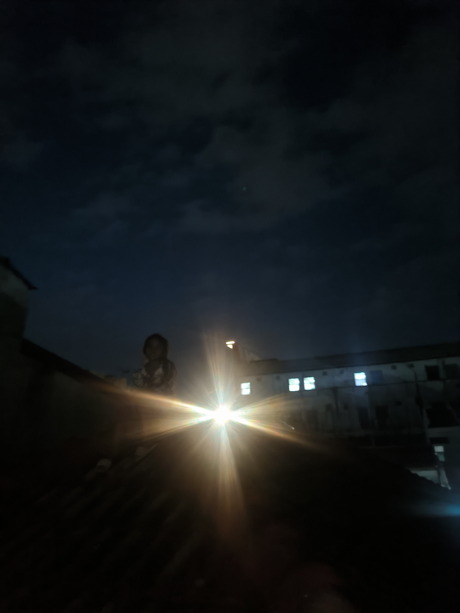 A nighttime image in Lhokseumawe, Indonesia. The image shows a person sitting on a dark roof, possibly made of corrugated metal. Behind the person is a bright, somewhat blurry, light source that casts a starburst effect, obscuring the view of what might be a building with multiple lit windows. The sky above is a cloudy, dark blue. The lower portion of the image is mostly dark with indistinct shapes.