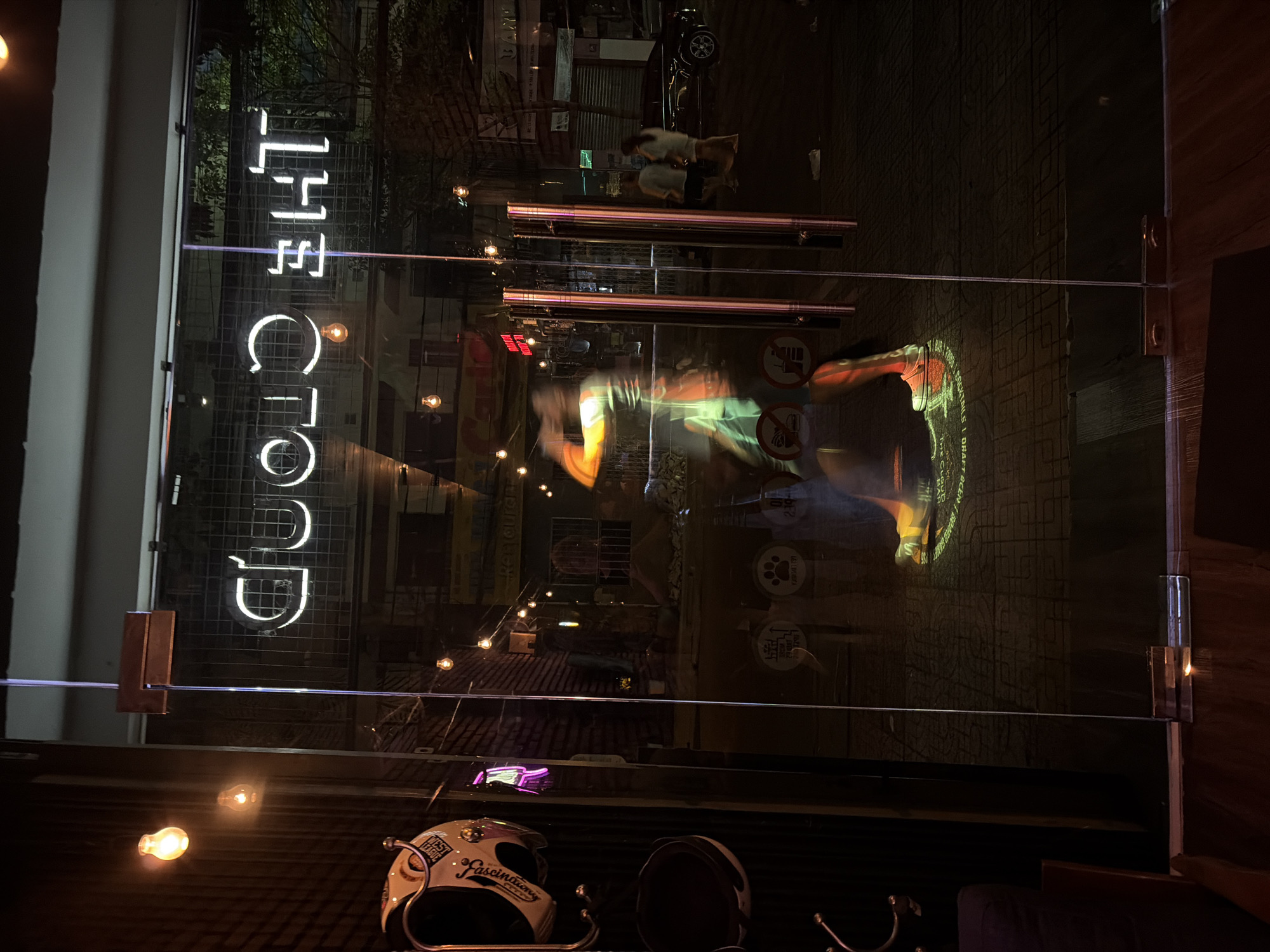 The image captures the entrance of a establishment at night, seen through a glass door. A neon sign spelling "THE CLOUD" is visible inside, reflecting on the glass. On the left, a white motorcycle helmet with colorful decals is hanging on a hook. Inside, several people are present. One person, blurred due to motion, appears to be walking or dancing, illuminated by a spotlight on the floor that projects a circular design. Two other individuals, a man and a woman, are walking outside on the street. The street is paved and dimly lit by streetlights and lights from buildings. In the background, shops and a parked car are visible. The overall scene suggests a vibrant nightlife setting in Nha Trang, Vietnam.
