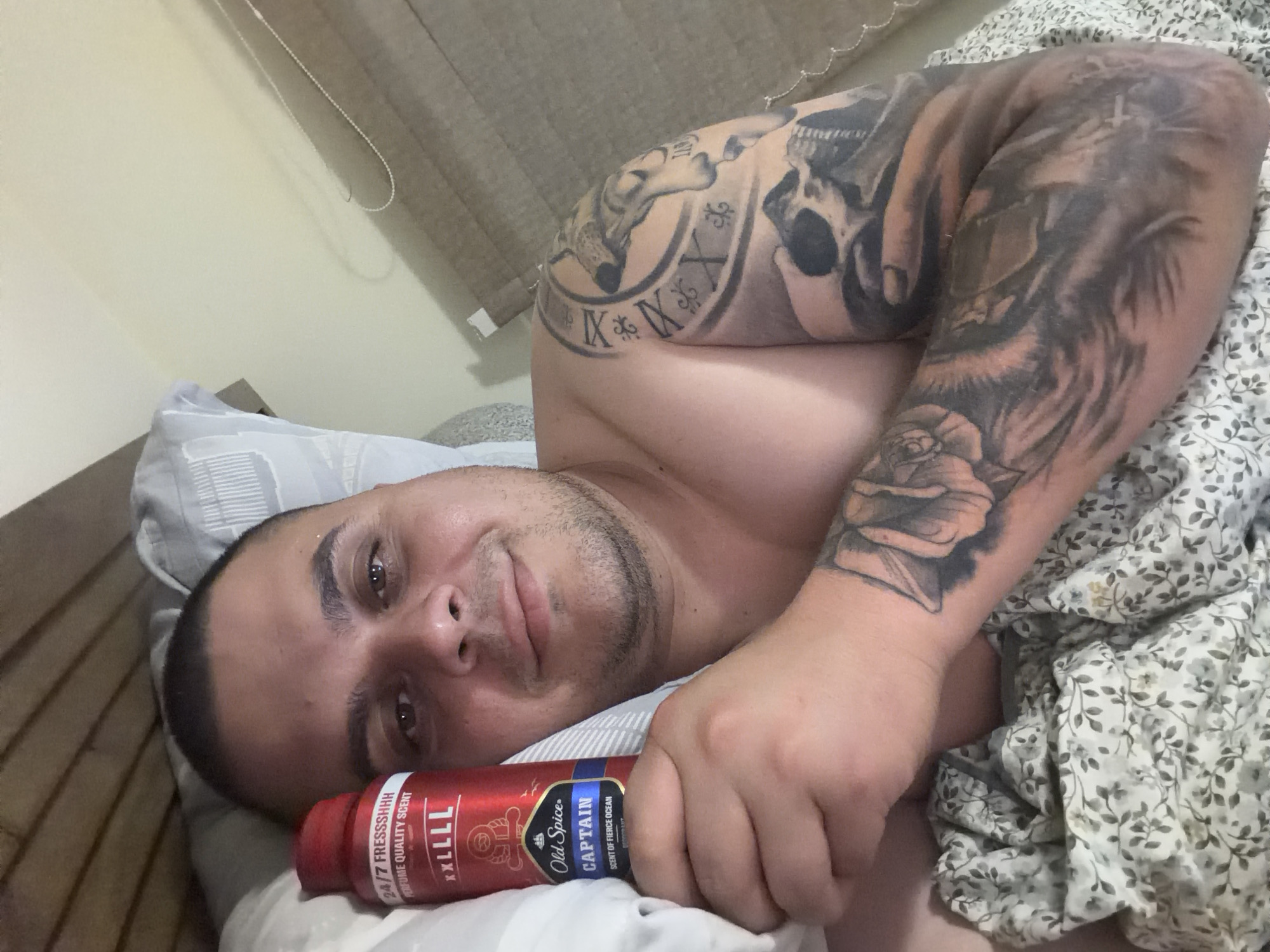 A man lies in bed, looking at the camera with a slight, relaxed smile. He has short dark hair and stubble, and his left arm is extensively tattooed with a skull, Roman numerals, a wolf-like figure, and a rose. He is shirtless and rests his head on a light-colored pillow. In his right hand, he holds a red Old Spice spray can. Visible text on the can includes "24/7 FRESSSHHH," "PERFUME QUALITY SCENT," "XXLLLL," "Old Spice," "CAPTAIN," and "SCENT OF FIERCE OCEAN." He is covered by a light-colored floral patterned blanket or duvet. In the background, a light-colored wall and a brown window blind are visible. The setting is a bedroom, suggesting a relaxed moment, and the location context is Shumen, Bulgaria.