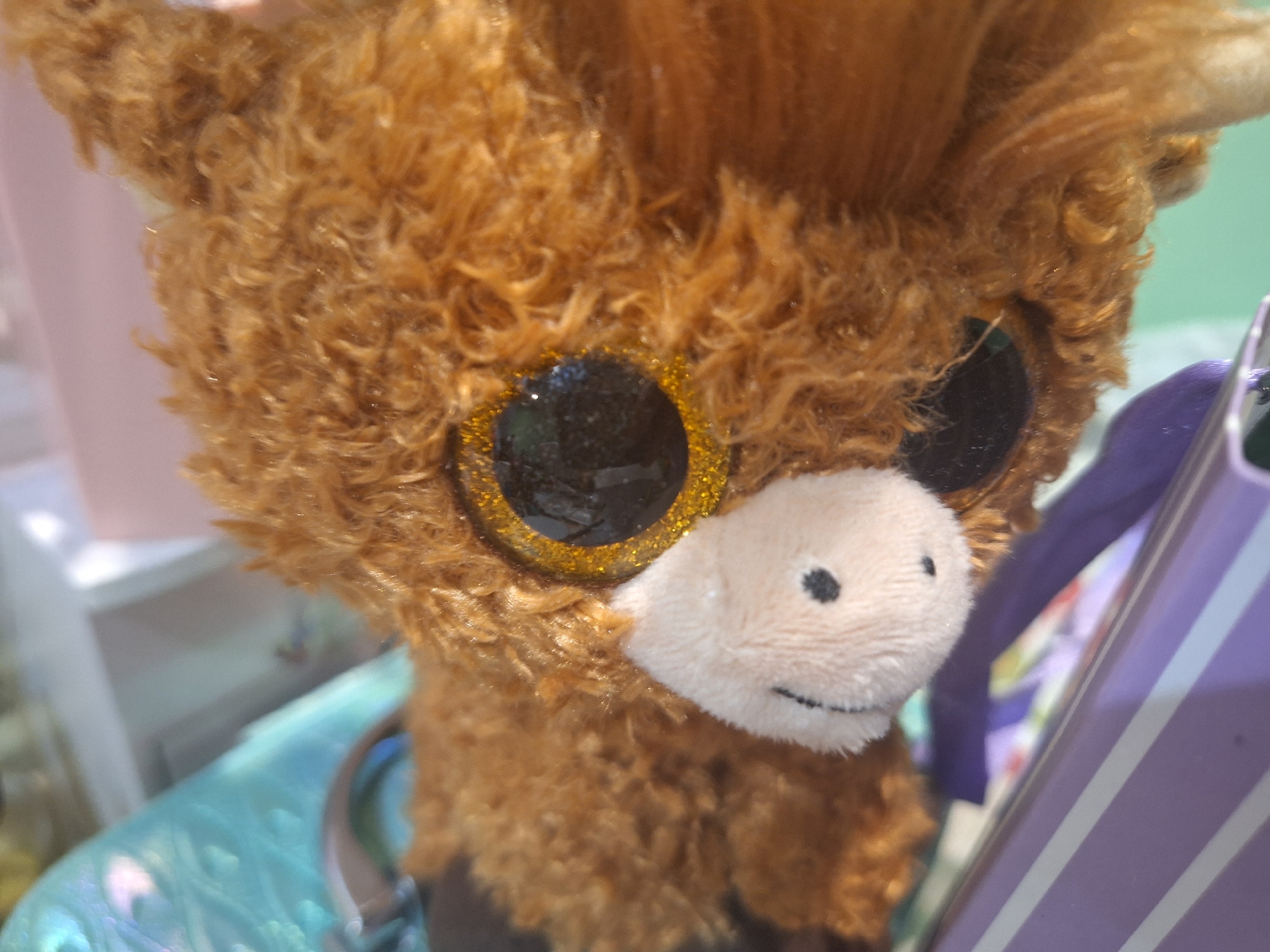 A close-up, eye-level, medium shot of a brown, shaggy, stuffed animal resembling a Highland cow. The stuffed animal's face is the primary focus, with large, glittery black eyes and a small black nose and smile on a light brown fabric muzzle. To the right of the stuffed animal is a purple and white striped object, possibly a box or container. In the background, a pale green surface is visible. The image appears to be taken indoors, possibly in a store or home environment. There is no indication of the city of Yambol, Bulgaria.