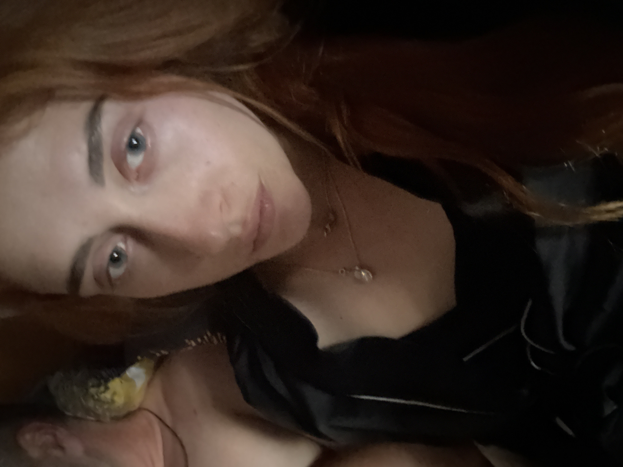 A selfie-style, low-angle shot features a woman with red hair and light blue eyes. She appears to be indoors, possibly in a private setting. She is wearing a dark-colored top with a white stripe, and a delicate necklace is visible around her neck. The location cannot be identified based on the provided image.
