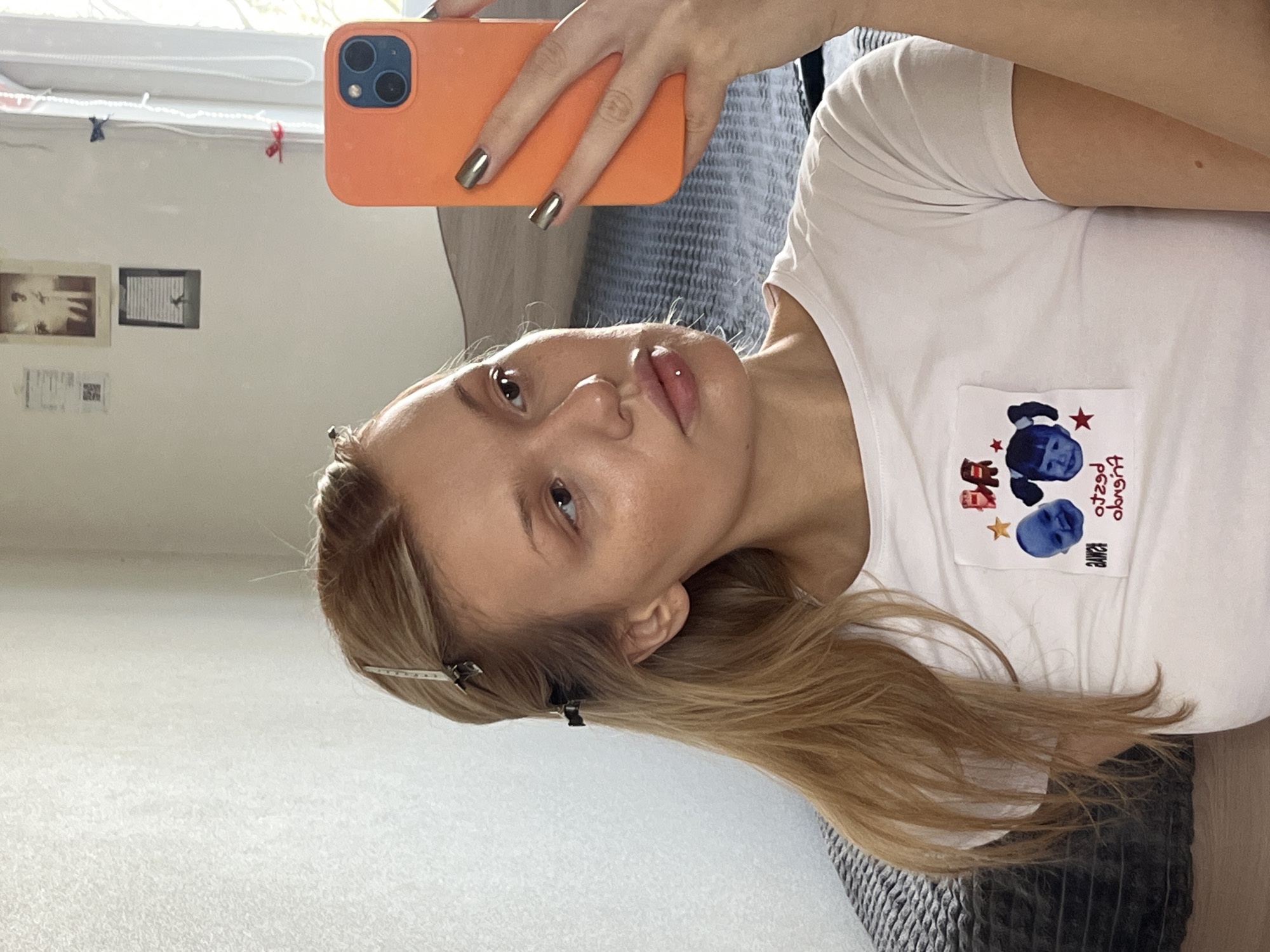 This media file is a mirror selfie featuring a young woman in an indoor setting, likely a bedroom or living room in Chaikovsky, Russia. The image is oriented with the subject's head to the right.
The central subject is a young woman with light skin, light-colored eyes, and long, light brown/blonde hair. Her hair is pulled back on the sides and held with small black clips, suggesting she might be styling it or letting it dry. She has a philtrum (medusa) piercing above her upper lip. Her fingernails are painted with a metallic dark silver or grey polish. She is holding an orange-cased smartphone, likely an iPhone given its visible camera module, to capture the image.
She is wearing a simple white t-shirt that features a small breast pocket. On this pocket, a colorful graphic design is visible, which appears reversed due to the mirror reflection. The graphic includes a small red figure (possibly a cartoon character), a yellow star, and two blue faces that resemble pop culture characters. Reversed text on the pocket can be partially deciphered as "p2sd origami" and "BANKS" or "BANKA".
The background, reflected in the mirror, reveals a light-colored wall illuminated by bright natural light streaming in from a window at the top. A string of white decorative lights is strung across the upper portion of the wall or window frame, adorned with small red and dark blue/black clips. Several framed items, including what appears to be an old black-and-white photograph, hang on the wall. A grey, textured fabric, likely a blanket or throw on a bed or sofa, is visible in the lower part of the reflection.
The bright lighting suggests it is daytime. The woman's expression is neutral as she looks directly into the phone/mirror. The overall scene depicts a casual and personal moment.