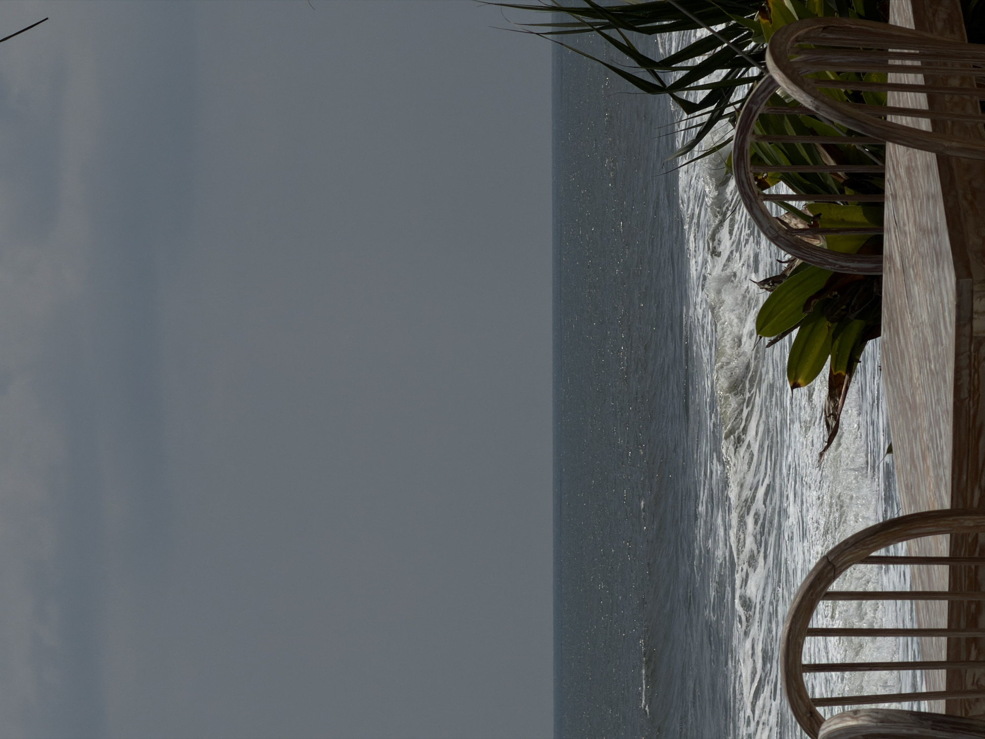 The image depicts an outdoor dining setup overlooking a turbulent ocean under an overcast sky in Belalang, Indonesia. In the immediate foreground, a light-toned wooden table is positioned with two matching slatted-back wooden chairs, oriented towards the water. Green tropical foliage, including broad leaves and spiky fronds, is visible between the furniture and the ocean, primarily on the right side. The ocean itself is dark blue-grey, characterized by multiple breaking waves that generate white foam. A distinct horizon line separates the ocean from a vast, dark grey, cloudy sky. No people or active events are present in the scene.