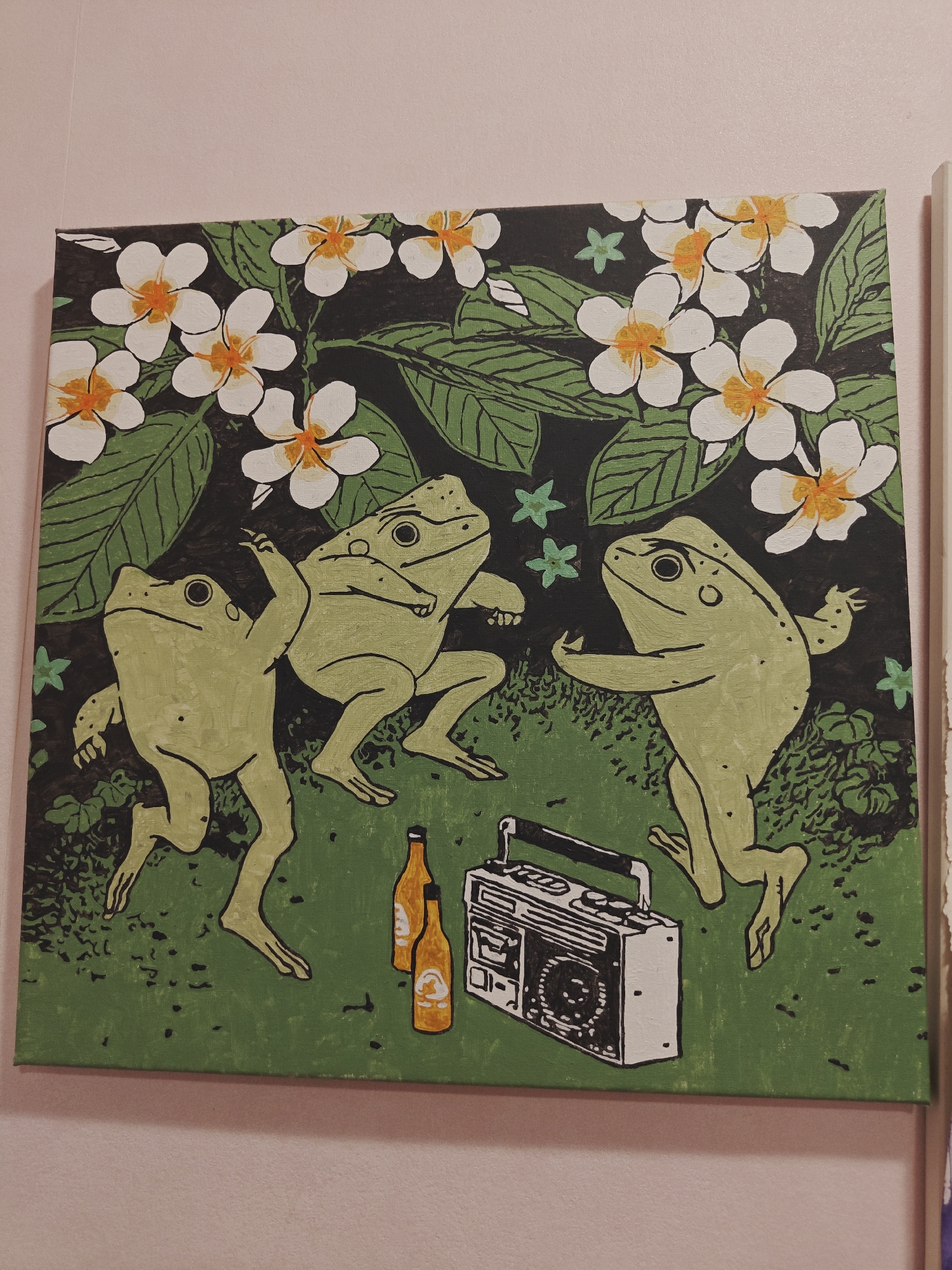 The image shows a painting on a canvas depicting three green frogs dancing on a grassy area. The frogs are stylized with simple features and are in various dynamic poses. Behind them, there are green leaves and white flowers with orange centers. In the foreground, there are two brown bottles and a black and white boombox. The painting is hanging on a light pink wall, possibly in a room in Saint Petersburg, Russia.