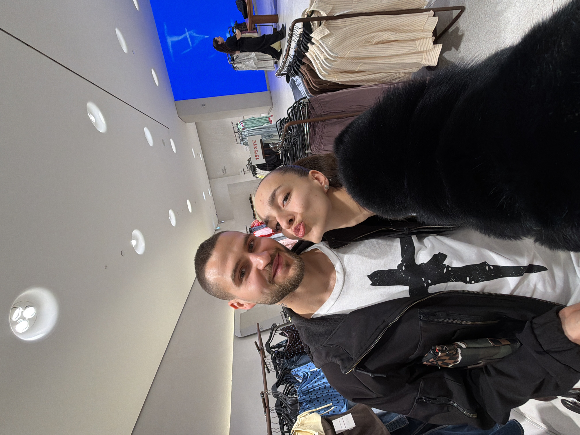 A selfie captures two individuals inside a modern, brightly lit retail clothing store, identified by Bulgarian signage as being in Sofia, Bulgaria, likely within a shopping mall. The male subject, positioned on the left, has short hair and a beard, wearing a black zip-up jacket over a white t-shirt featuring a large black, stylized cross graphic. He smiles subtly at the camera. The female subject, on the right and partially obscured, has dark hair pulled back and is making a kissing gesture towards the camera. She wears a black, presumably faux fur, garment and a silver earring.
In the mid-ground, a white sign clearly displays the Bulgarian text "КАСА ПРОБА," indicating a checkout or fitting room area. Another smaller white sign shows "15M/31??". The store features numerous clothing racks laden with various garments, including patterned tops, denim, and striped shirts. A large, prominent blue digital display screen showing abstract patterns is visible in the background on the right. A third female, appearing to be a store employee in dark attire, walks from right to left in the background near the blue screen. The store's interior features a white ceiling with multiple recessed circular lights and a light-colored floor. The male subject has a small, checkered-patterned object tucked into a pocket of his jacket.