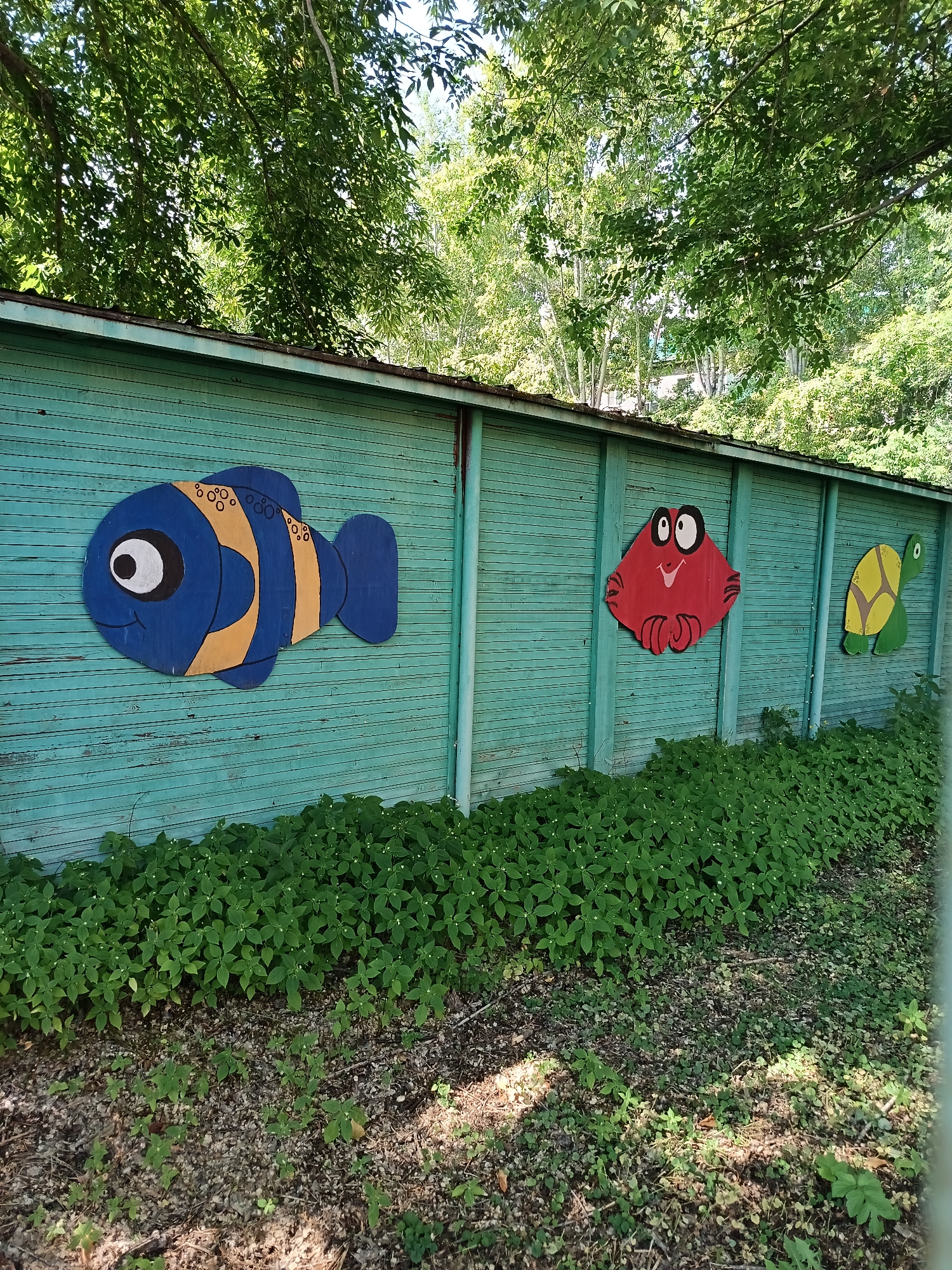 The image depicts a light blue wooden fence with three painted wooden marine animals affixed to it: a blue and yellow fish, a red crab, and a green and yellow turtle. The fence is located outdoors, with lush green foliage growing at its base. Overhead, the branches of a large tree with green leaves obscure the sky, suggesting a natural or park-like setting. The fence appears weathered, with visible horizontal planks. The style of the artwork suggests a children's play area or a decorative outdoor feature. The image does not contain any individuals. The specific location within Komsomolsk-on-Amur, Russia, cannot be determined from the visual information provided.