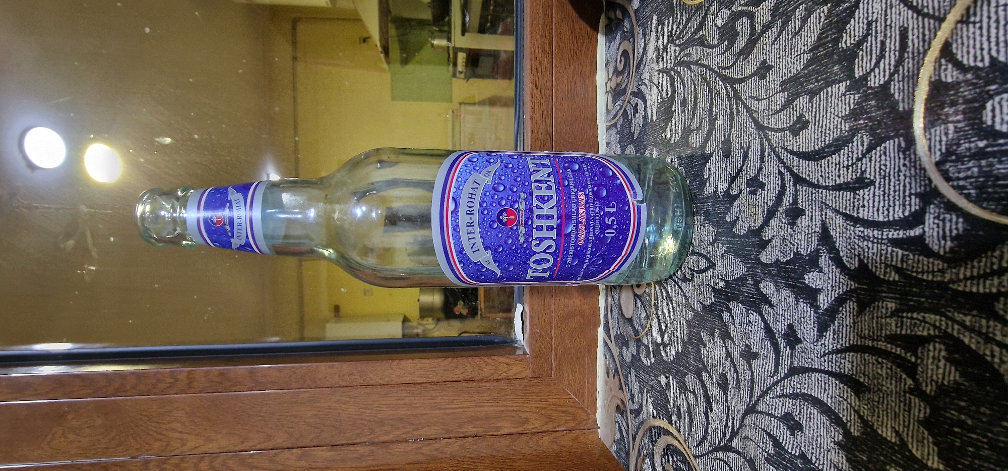 A clear glass bottle of Tashkent sparkling water is positioned centrally on a patterned surface in front of a reflective surface that shows a kitchen scene. The bottle has a blue label with white text, including "INTER-ROHAT" and "TOSHKENT," and a volume of "0.5 L". The reflective surface behind the bottle shows kitchen cabinets, a stove, and lighting fixtures. The patterned surface is dark with lighter swirling designs. The scene appears to be indoors, possibly in Karshi City, Uzbekistan, given the brand of the water.