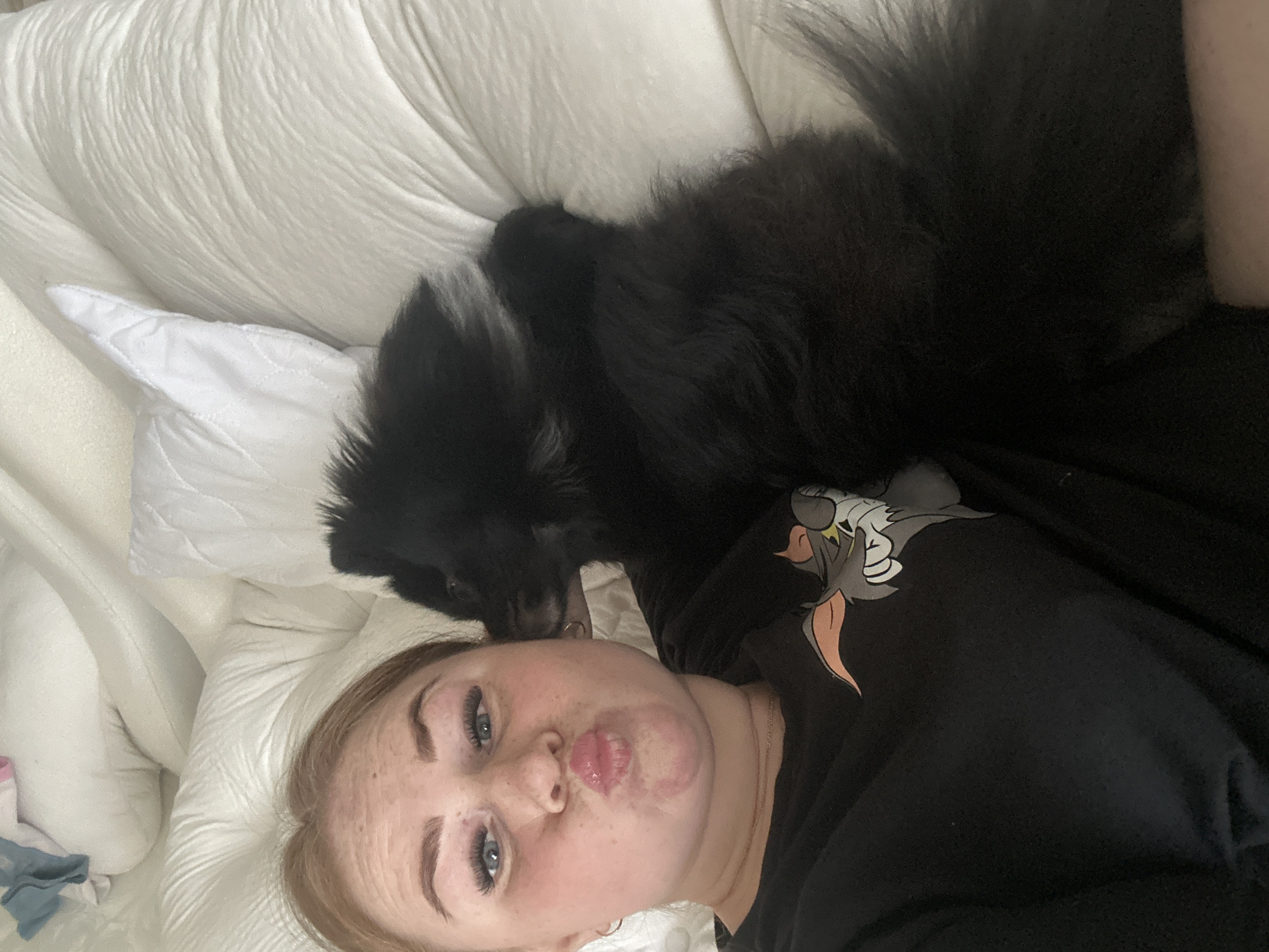 An overhead-oriented photograph captures a young adult female lying on her left side, partially facing the camera, alongside a black dog. The female has light-colored hair, a fair complexion with visible freckles, blue eyes, and distinct dark eyelashes and eyebrows. Her lips are puckered into a "kiss" expression, with faint red markings visible around her mouth. She wears a black t-shirt featuring a graphic of the cartoon character Tom from "Tom and Jerry" on the left chest area and a thin gold chain around her neck. The black dog, which appears to be a long-haired breed with some white markings on its chest, is positioned to her right, resting its head near her shoulder/neck. Both subjects are on a bed or sofa covered with white, quilted bedding, including a pillow beneath the female's head. In the upper right background, crumpled light blue and white fabric items are visible on the bedding. The setting is indoors. The specific location within the city of Chaikovsky, Russia, cannot be determined from the provided image.