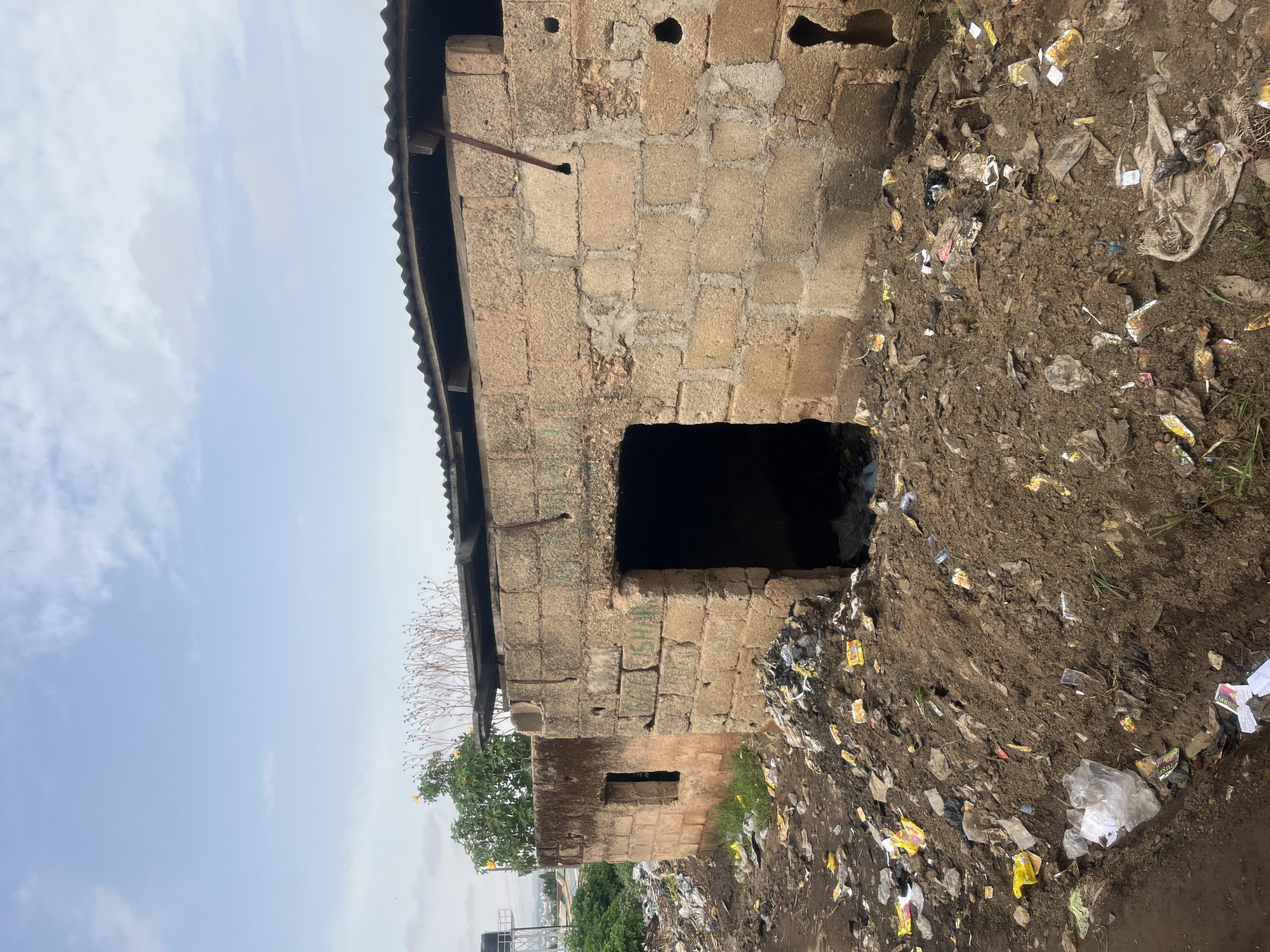 The image depicts a dilapidated, unfinished block building in Gura Top, Nigeria. The structure is constructed from roughly hewn concrete blocks. The building appears to be abandoned; the ground surrounding the structure is covered with garbage. The sky is partly cloudy.